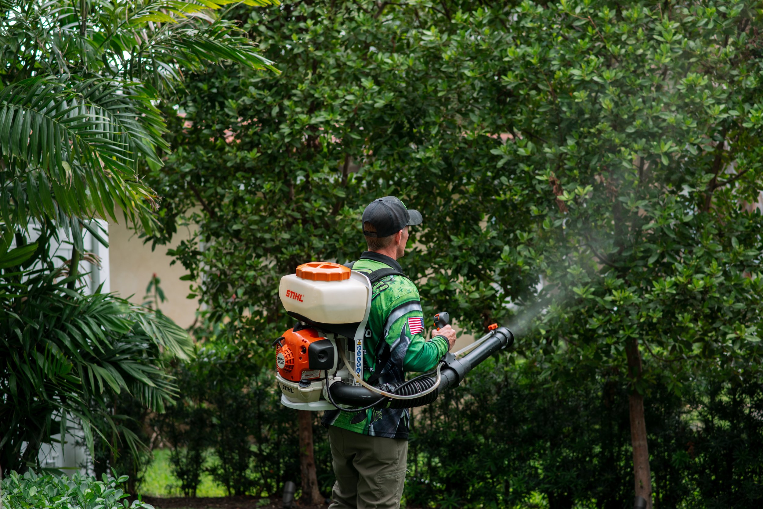 A man wearing a green and black shirt, khaki pants, and a cap is using a backpack leaf blower to trim bushes and plants in a garden surrounded by lush green trees and foliage.