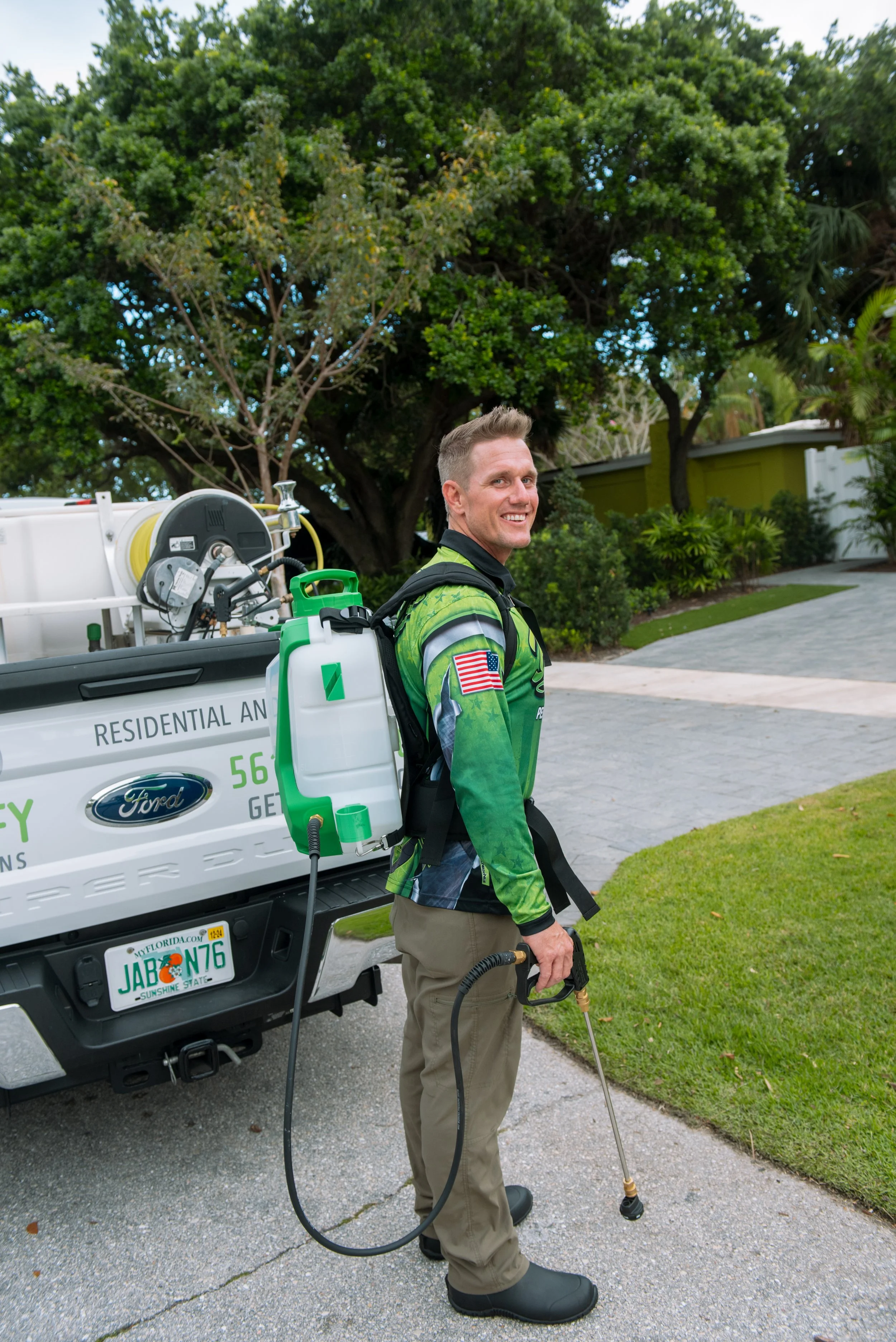 A smiling man wearing a green and black uniform with an American flag patch on the shoulder, standing on a driveway, holding a pest control sprayer wand, with a fleet of pest control equipment mounted on a white Ford truck behind him, in front of a large tree and residential house.