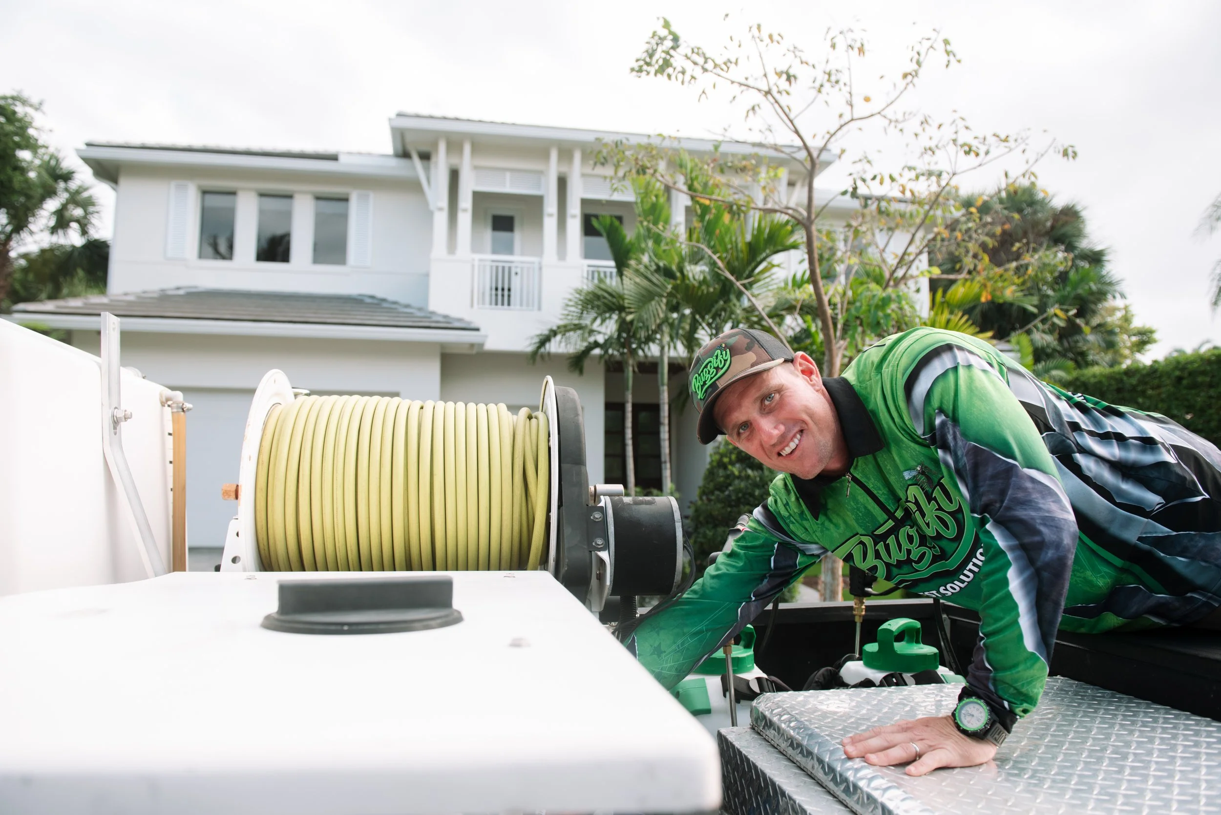 A man wearing a green and black cycling jersey and a camouflage cap working on equipment on a roof in front of a modern white house with trees and cloudy sky in the background.