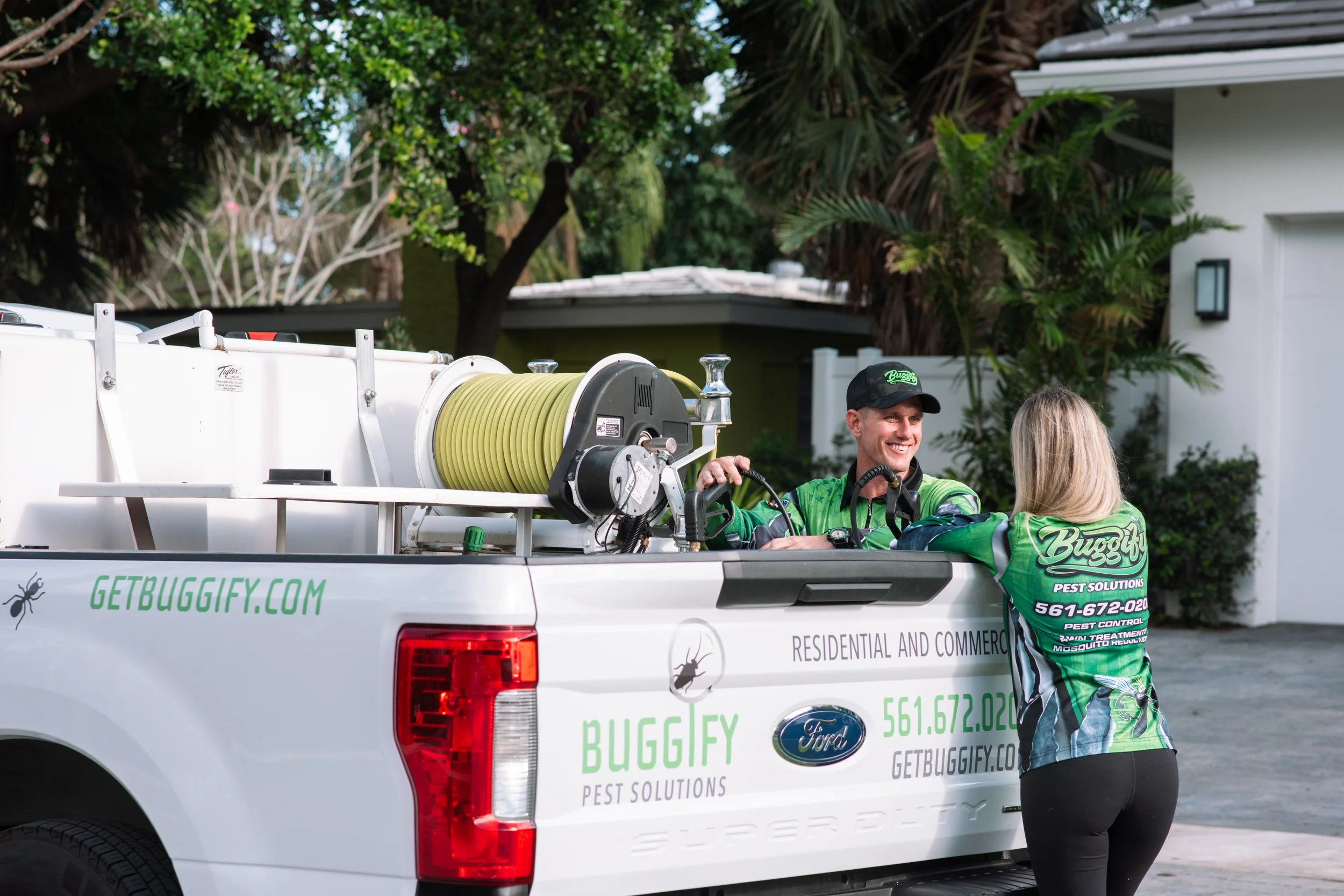 Two pest control workers in green uniforms standing on the back of a white truck, smiling and talking to each other. The truck has the words GETBUGGIFY.COM and pest control contact info printed on it, along with a logo and a picture of a bug.