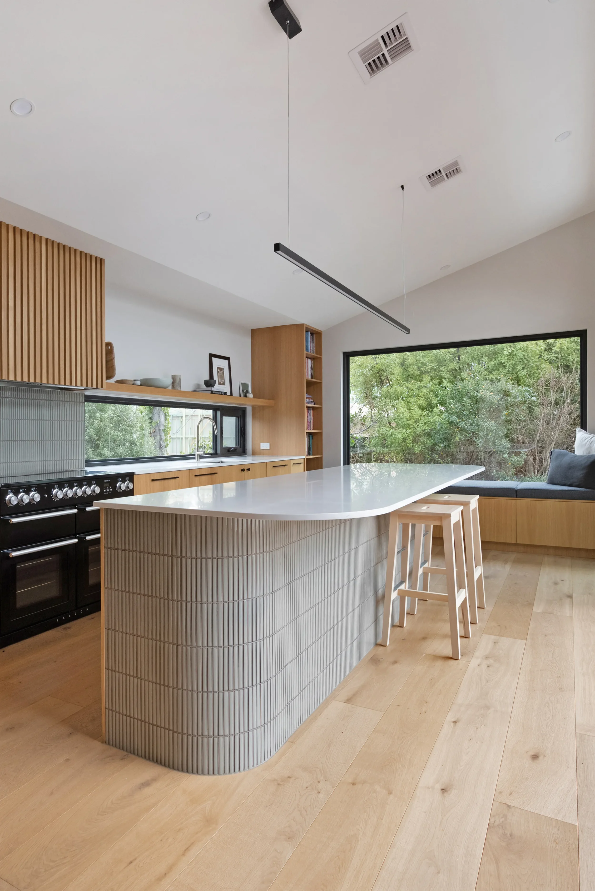 Modern kitchen with a large white island, wooden stools, a black oven, and built-in wooden shelves. Large windows overlook green trees outside. Ceiling has modern lighting fixtures and air vents.