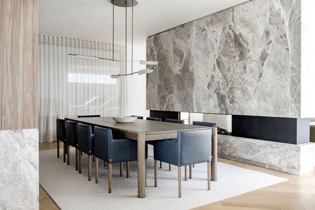 Modern dining room with a large wooden table, eight navy blue chairs, a marble fireplace, light wood flooring, and a window with white curtains.