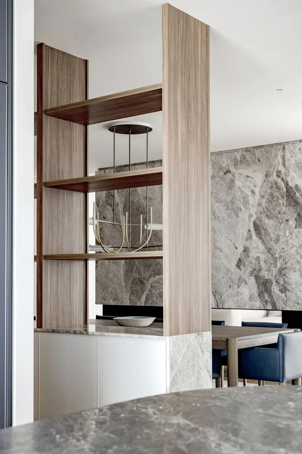 Modern interior with wooden open shelves, a gold chandelier, marble accents, and a dining area with blue chairs.