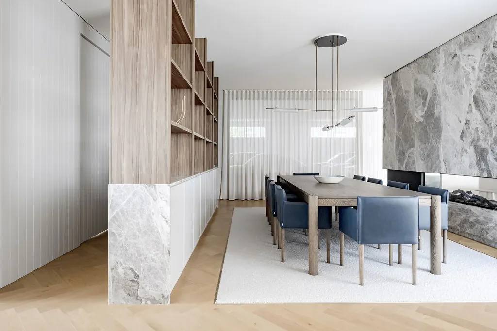 Modern dining room with wooden table, blue upholstered chairs, a marble fireplace, a large wooden bookshelf, white curtains, and minimalist contemporary decor.