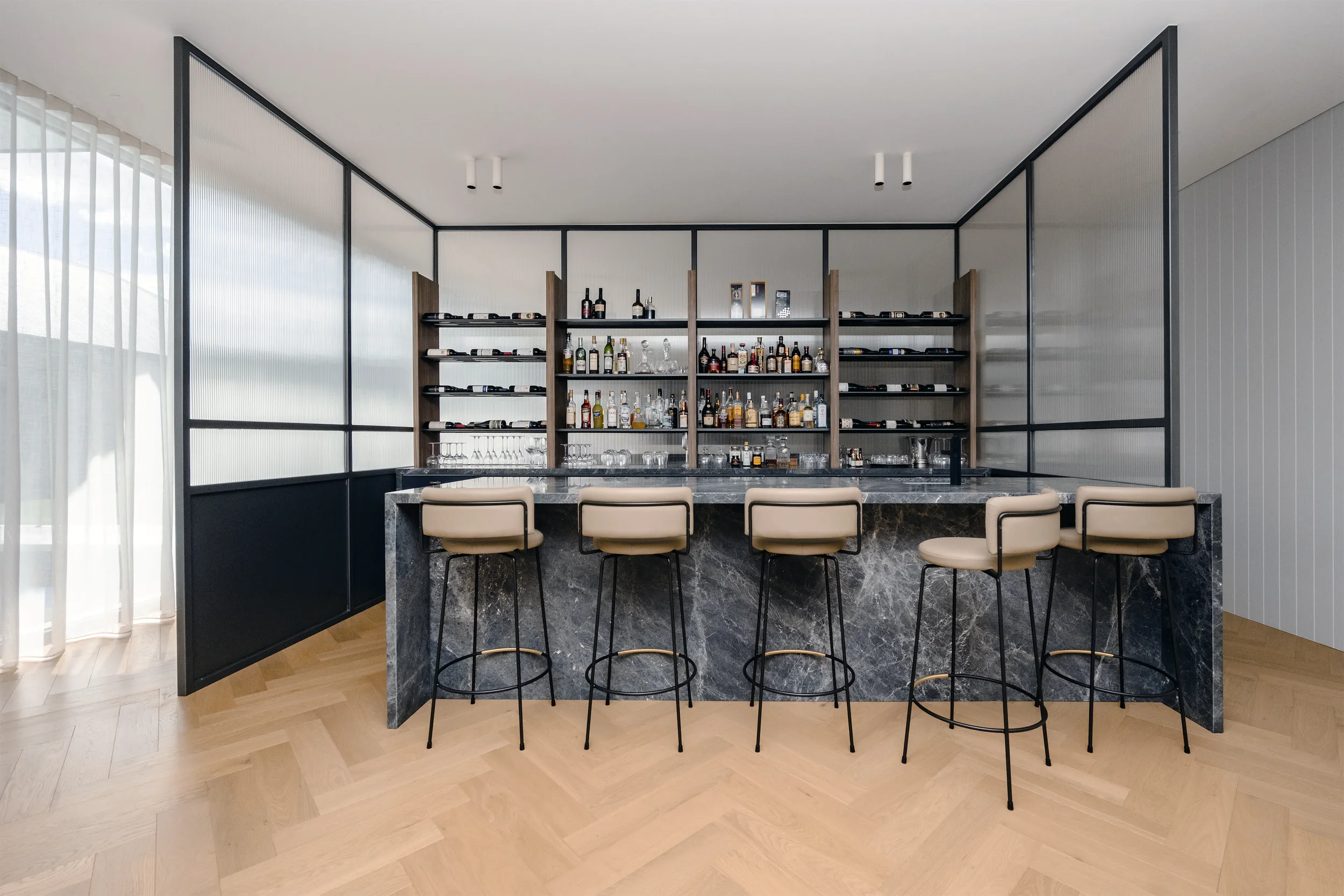 A modern bar with a black marble countertop, white upholstered bar stools, and open shelving behind with bottles of alcohol and glassware. Large windows with sheer curtains allow natural light into the space.