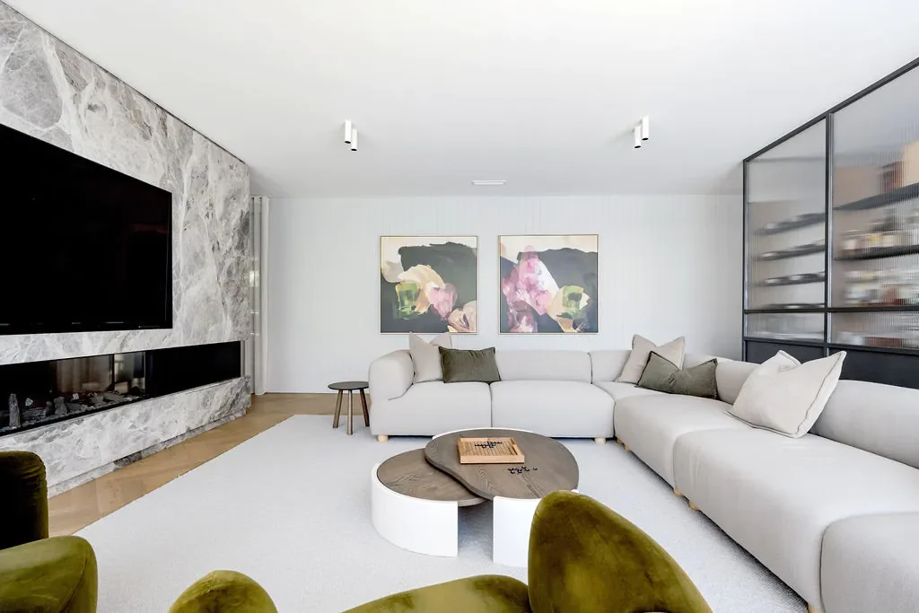 Modern living room with a large L-shaped cream-colored sofa, abstract paintings on the wall, a stone fireplace, a flat-screen TV, a glass-paneled wall, and a round coffee table.