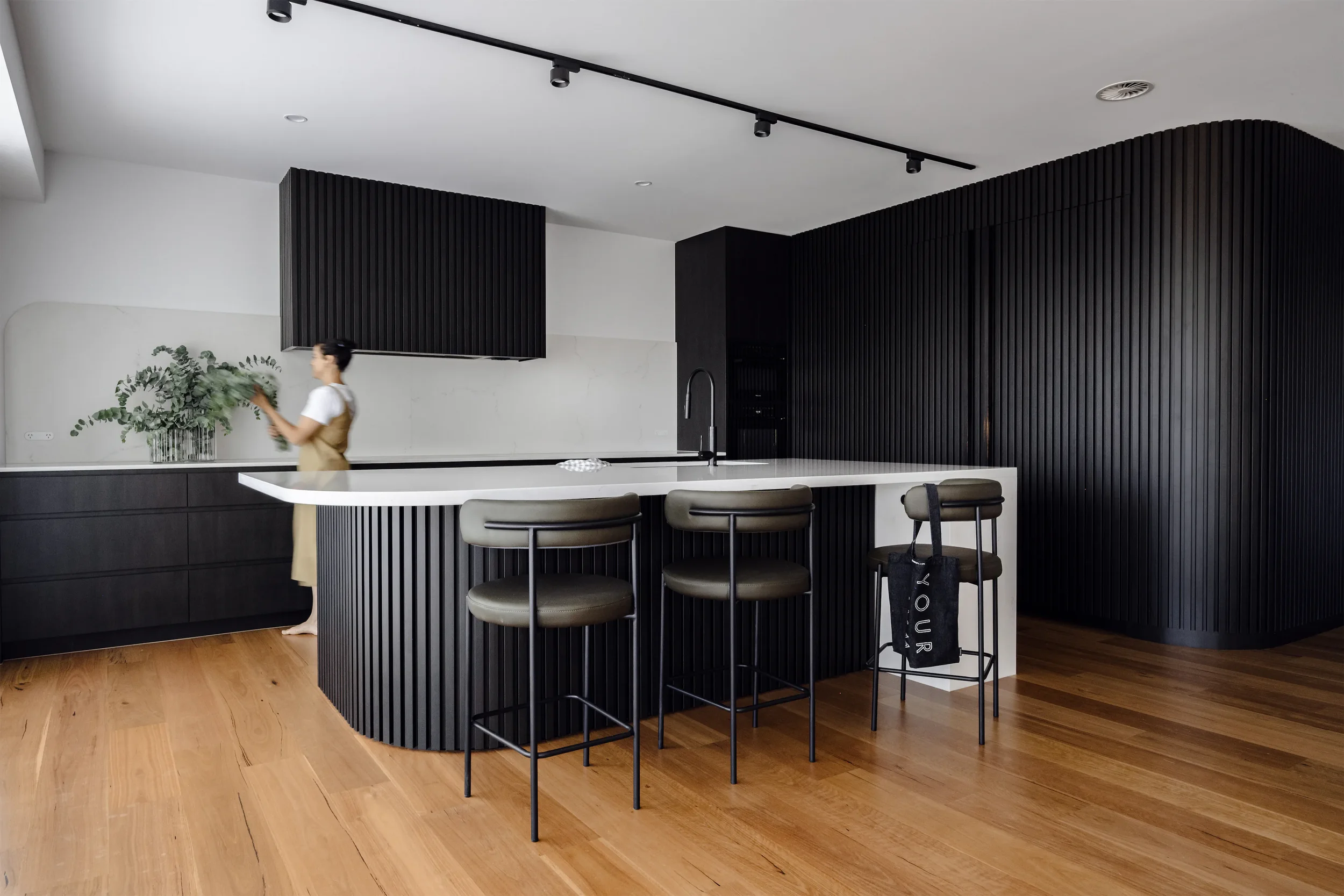 A modern kitchen with a white countertop, black vertical slatted panels, and a black track lighting on a white ceiling. A woman is arranging a plant on the counter, with a black bag hanging from a stool.