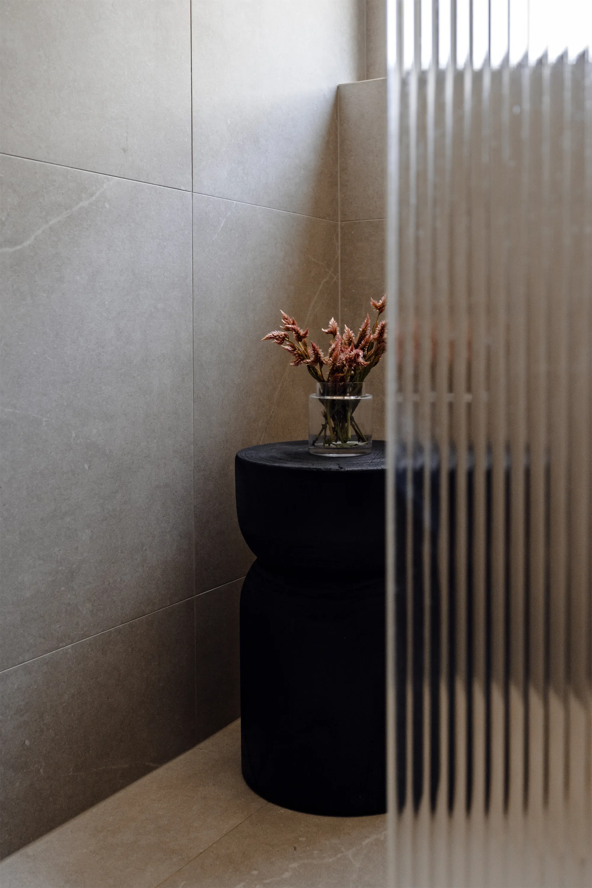 A black toilet with a glass vase containing pink curly flowers on top, placed on a black surface in a bathroom with beige tiled walls and a partial view of a white ribbed partition.