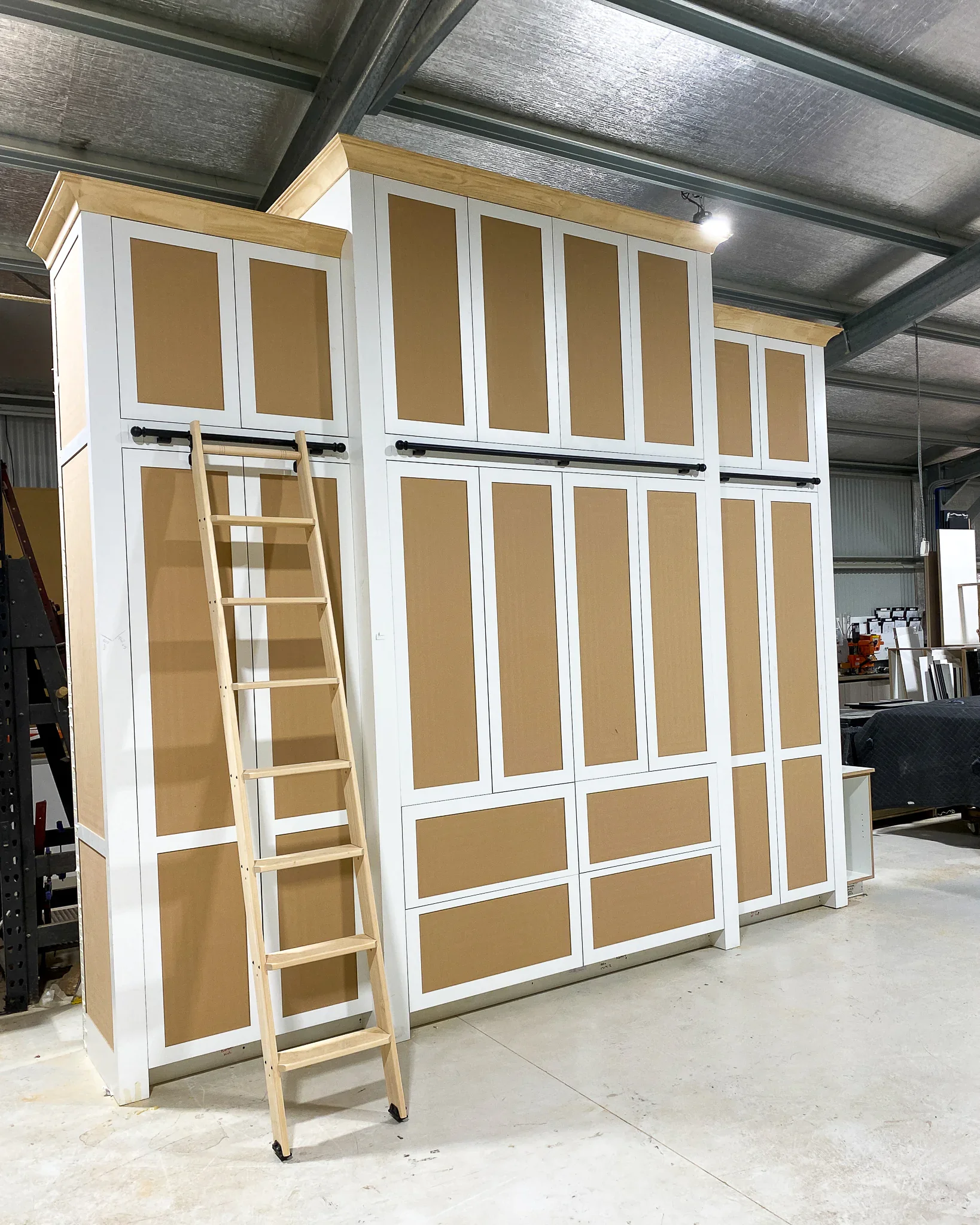 A built-in unit with white framing and brown paneling, featuring two rows of cabinet doors. A small wooden stepladder is leaning against the unit. The setting appears to be a warehouse or workshop with metal roofing and additional stored items visible in the background.