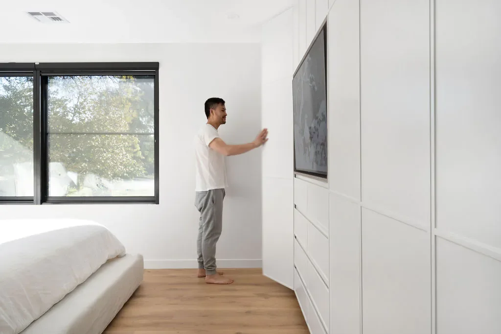 A man standing barefoot in a bedroom, touching a white wall next to a mounted TV, with a bed and a window showing trees outside.