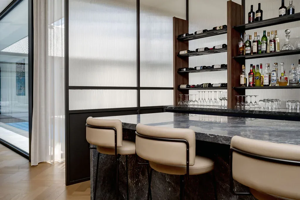 Modern home bar with a marble countertop, beige stools, and open shelves stocked with liquor bottles and glassware