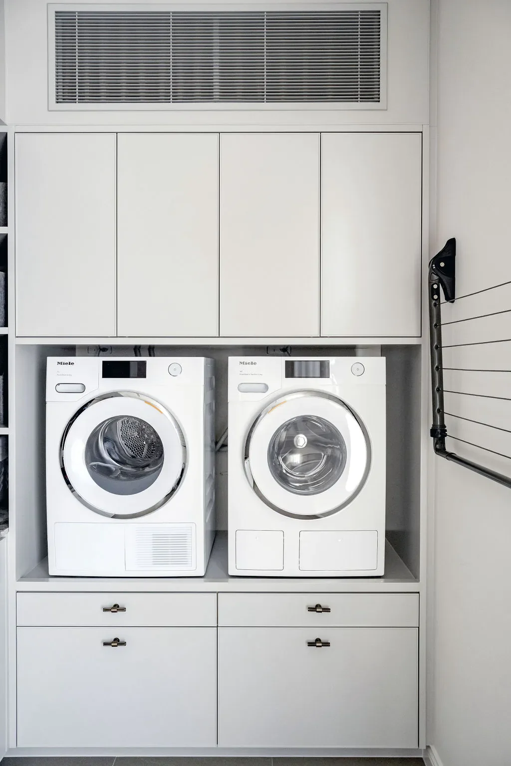 White front-loading washer and dryer side by side in a laundry room with build-in white cabinetry above and below, a vent on the wall to the right, and a black broom handle leaning against the cabinetry.