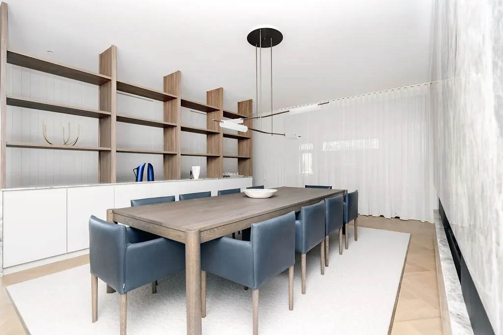 Modern dining room with a wooden table, eight blue chairs, a white rug, and built-in shelving on the wall with decorative items.