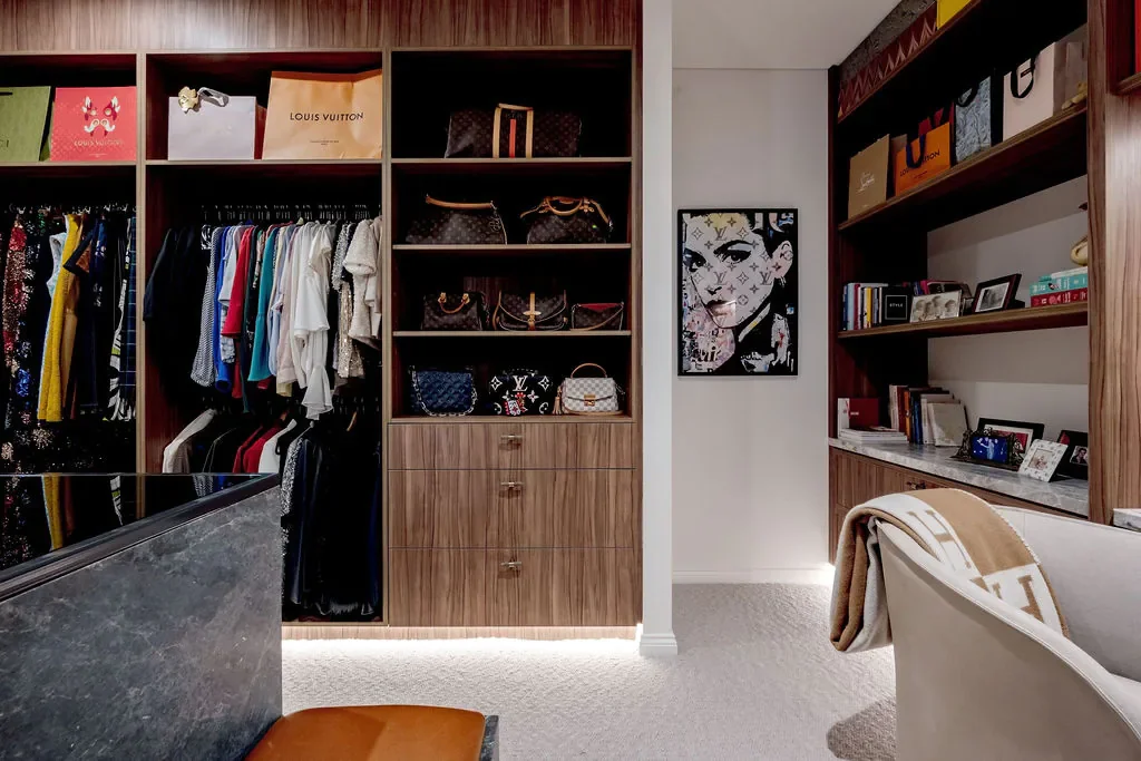 Luxury closet with shelves of handbags, clothing racks with colorful clothes, a framed artwork of a woman's face, and a bookshelf with framed photos and books.