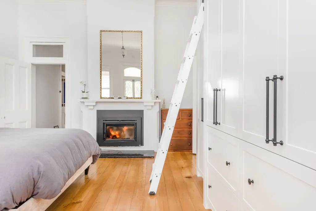 Interior view of a bedroom with a fireplace, a ladder, a bed with gray bedding, large white cabinets, and a mirror above the fireplace reflecting a window.