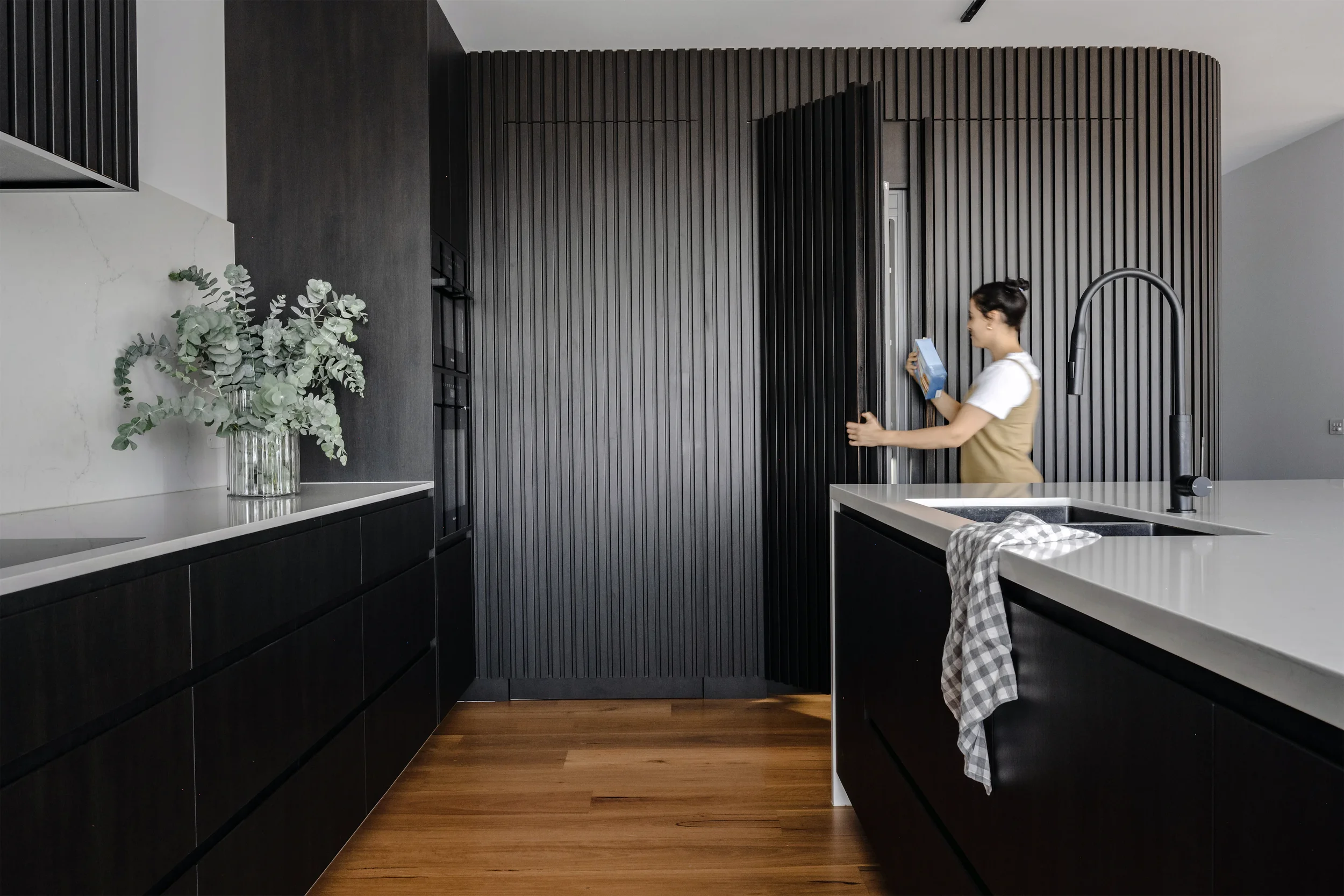 Modern kitchen with black cabinetry, wooden flooring, a white countertop, a vase with green and white leaves, and a woman opening a black cabinet door while holding a box and wearing a white shirt and beige vest.