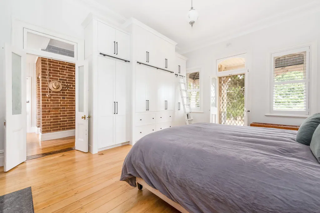 Bright bedroom with white walls, hardwood floors, a large bed with gray bedding, and a built-in white closet. There are two windows with blinds and a door leading outside, with a ladder near the closet.
