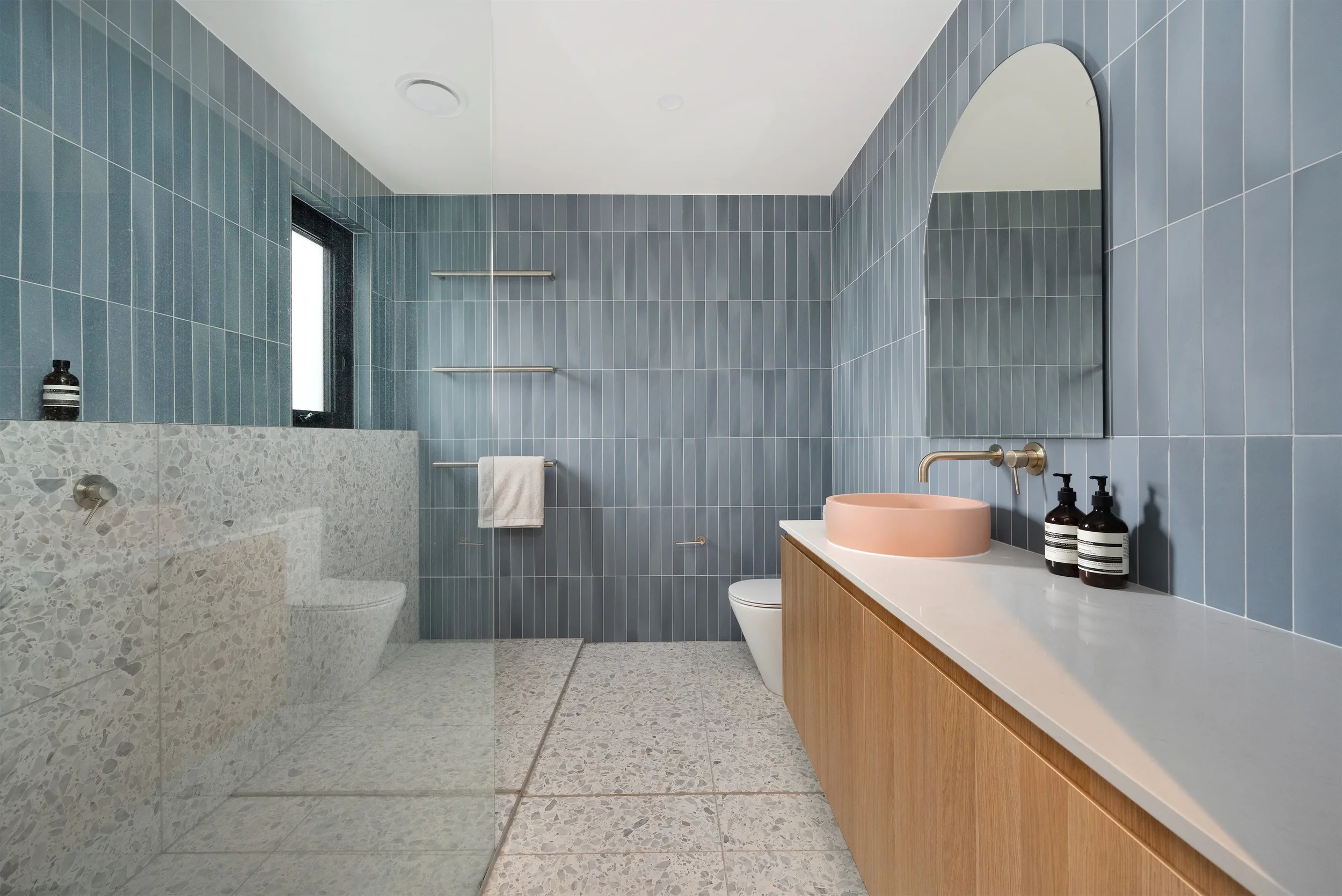 Modern bathroom with blue vertical tile walls, terrazzo countertop with a pink vessel sink, wooden vanity, large mirror, and black soap dispensers.