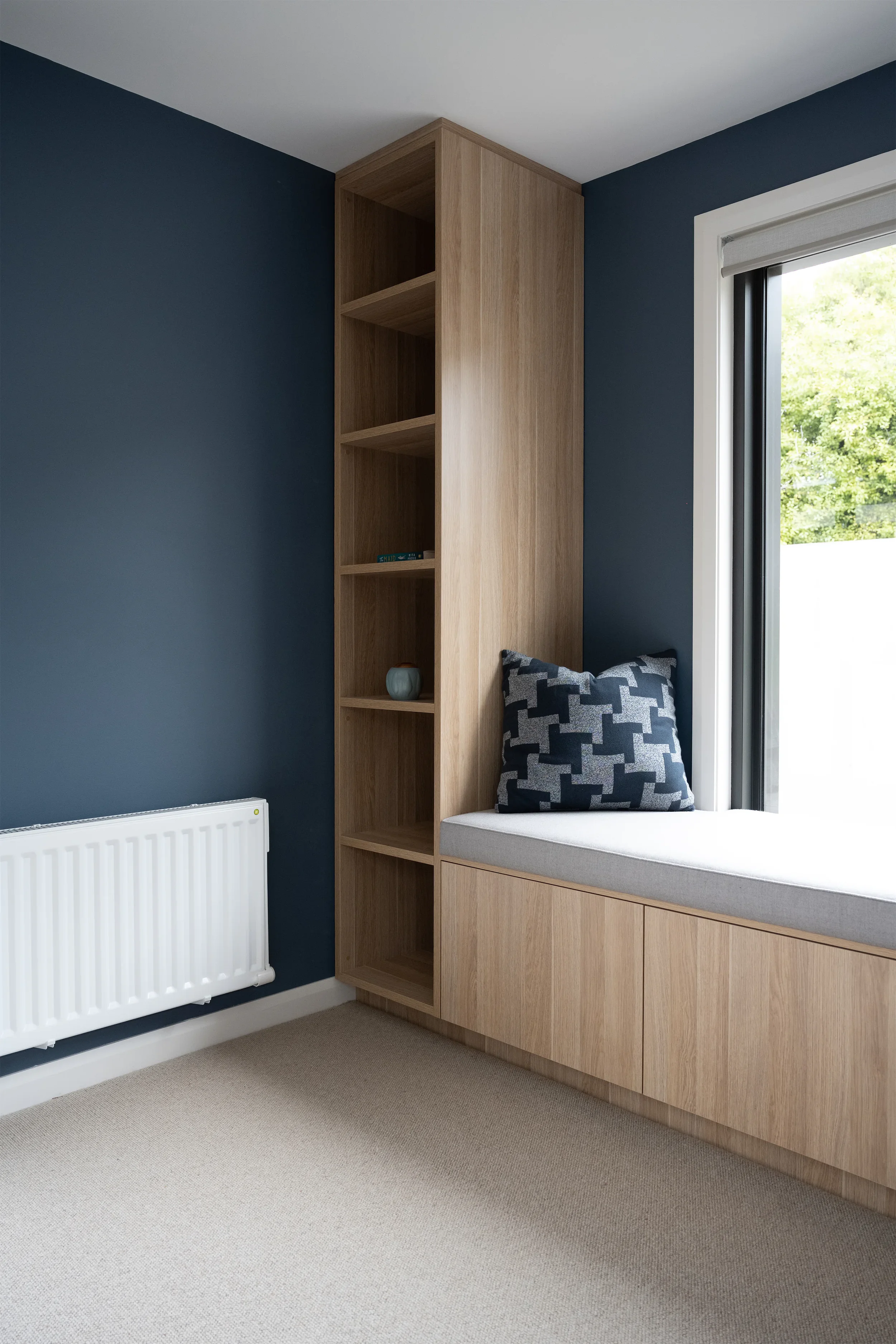 A corner of a room with a navy blue wall, a built-in wooden shelving unit, and a window with a view of green trees. There is a beige cushioned window seat with a patterned pillow, a white radiator, and beige carpeting.