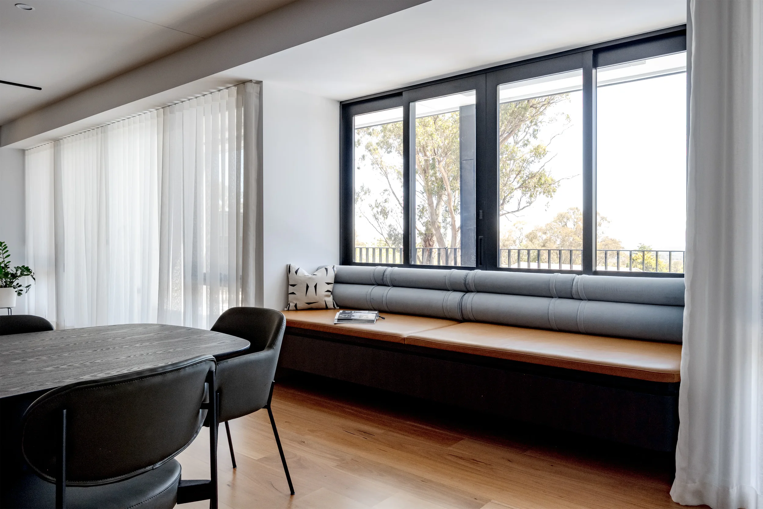 Living room with large windows, white curtains, a white cushioned window seat with a patterned pillow, a round wooden table, black chairs, and a potted plant, overlooking trees outside.