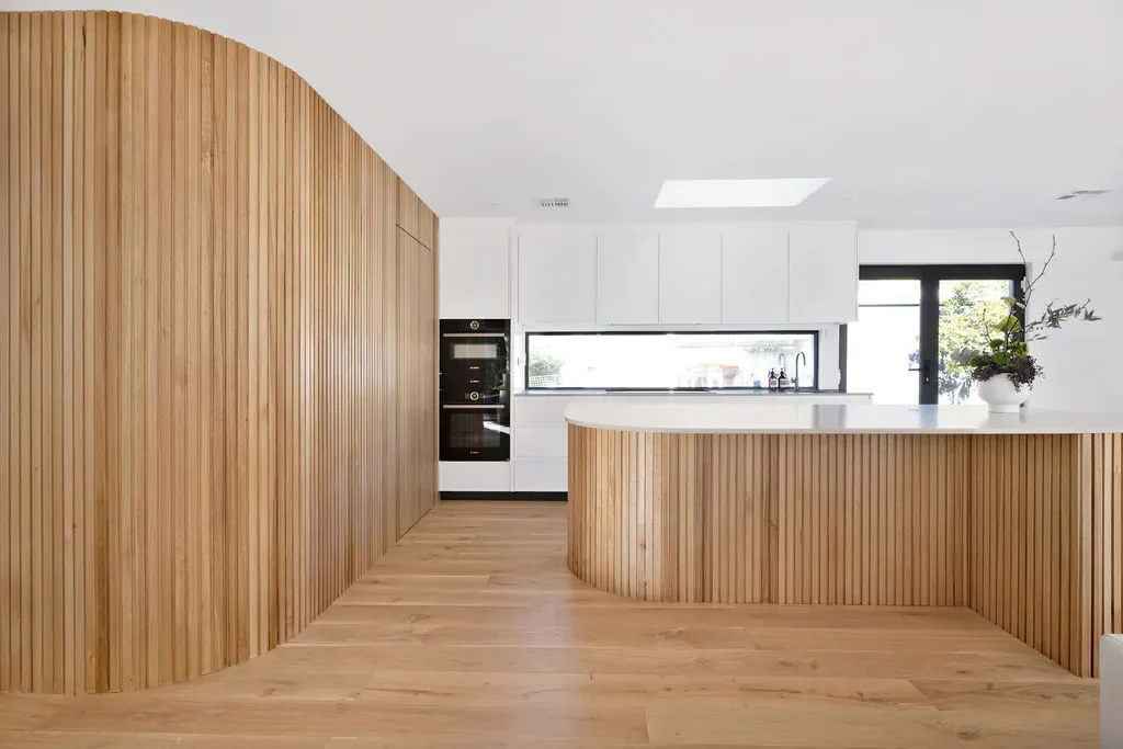 Modern kitchen with wooden cabinetry and flooring, white countertops, built-in black oven, large window, and decorative plant in a white vase.