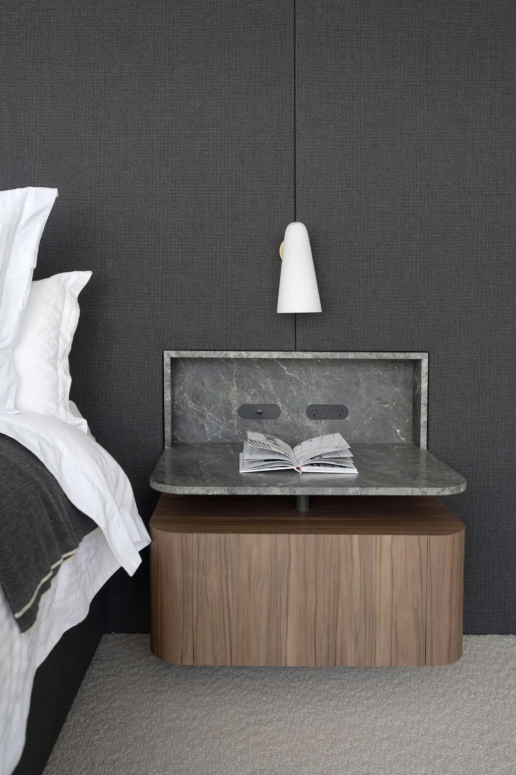 A bedside table with a marble top, an open book, and two electrical outlets, mounted against a dark textured wall with a white wall sconce hanging above.