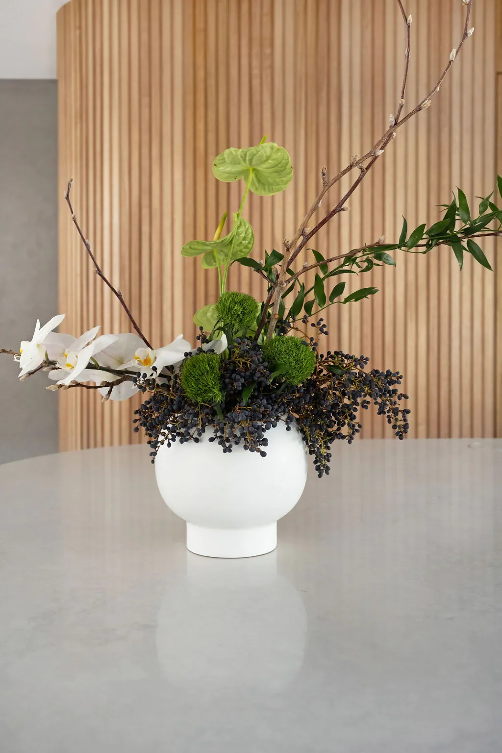 A white vase with mixed green and white flowers, branches, and dark berries on a light gray table, with a wooden slatted wall in the background.