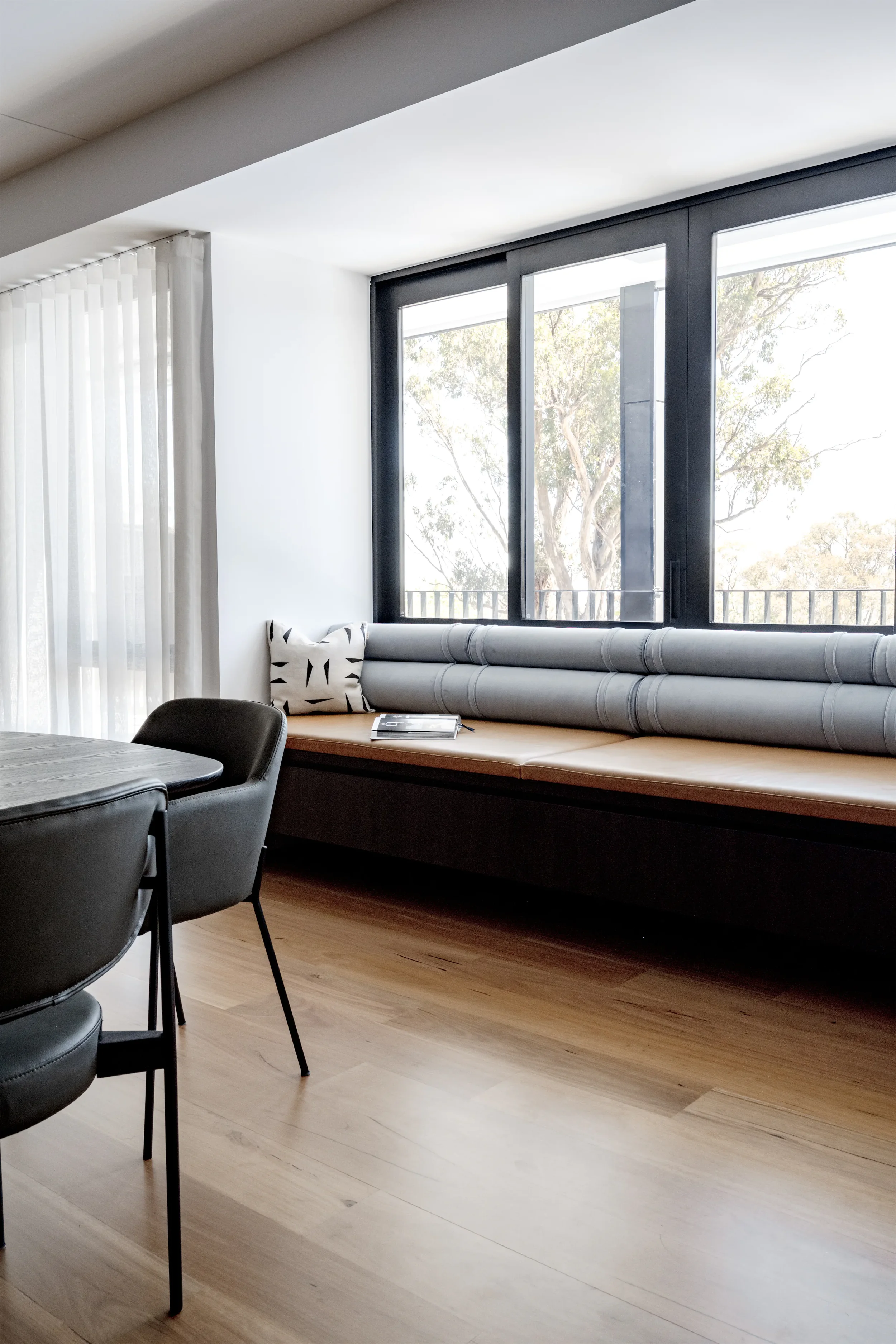 Modern living room with large windows showing trees outside, a built-in window seat with cushions, a table with magazines, black chairs, white curtains, and hardwood flooring.