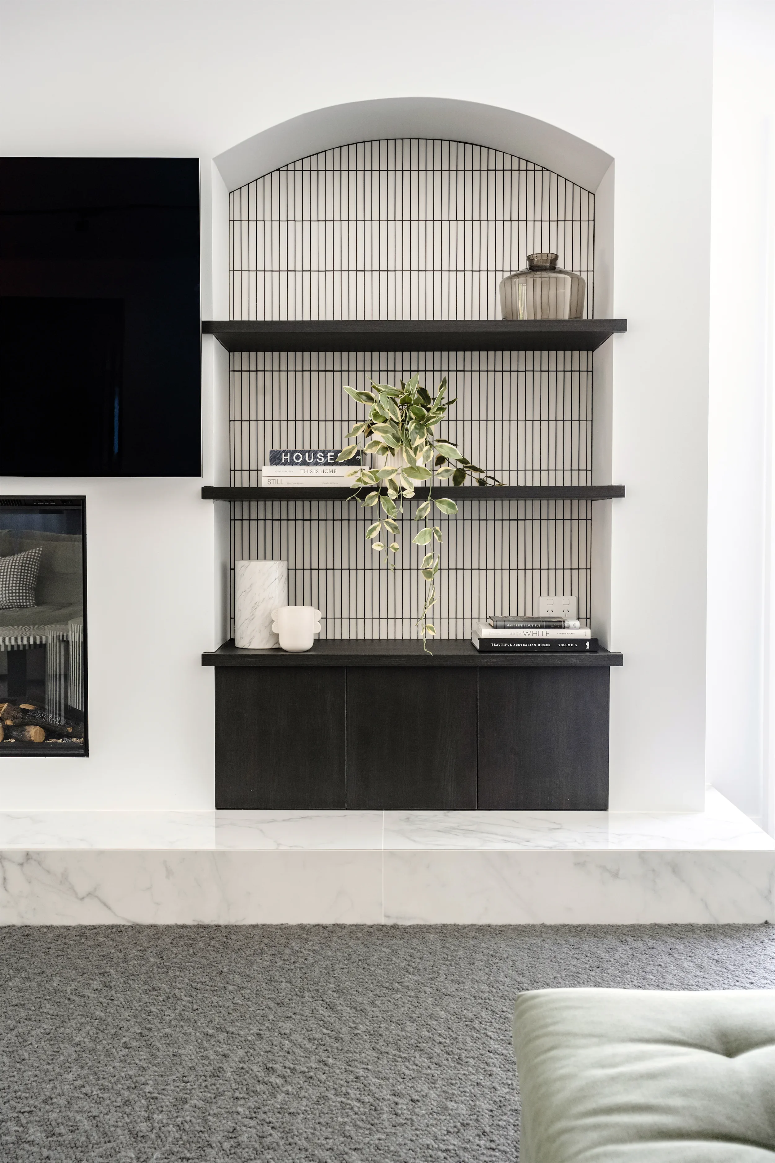 Living room with a black entertainment unit on a wall featuring black wire shelving, decorative books, a white vase, and a potted plant on the shelves. The room has white walls, a marble-like baseboard, and a gray carpeted floor.