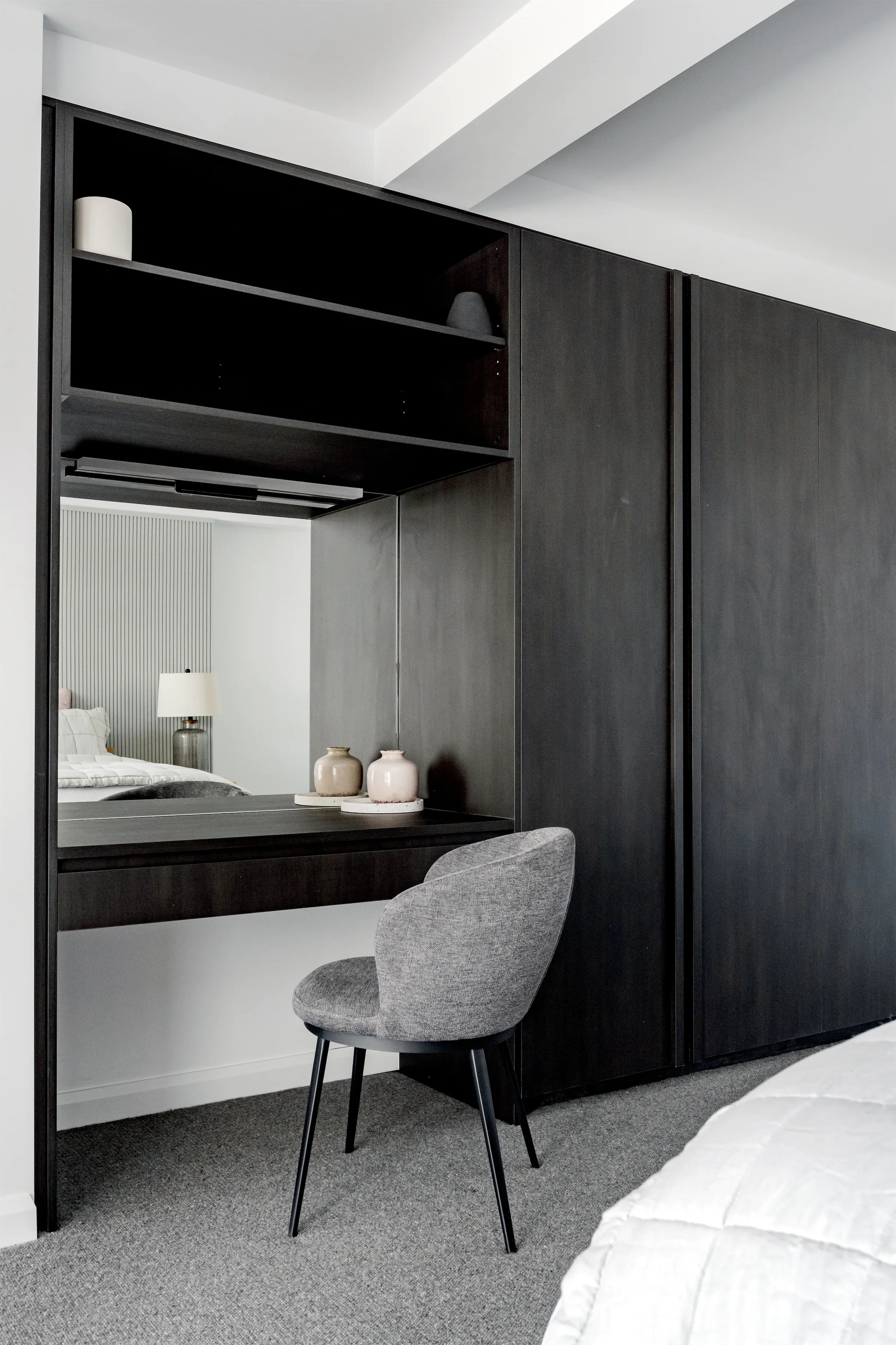 A modern bedroom with a black wardrobe, a built-in desk with vases, a gray upholstered chair, and a bed with white bedding. A mirror reflects a bedside lamp and part of the bed.