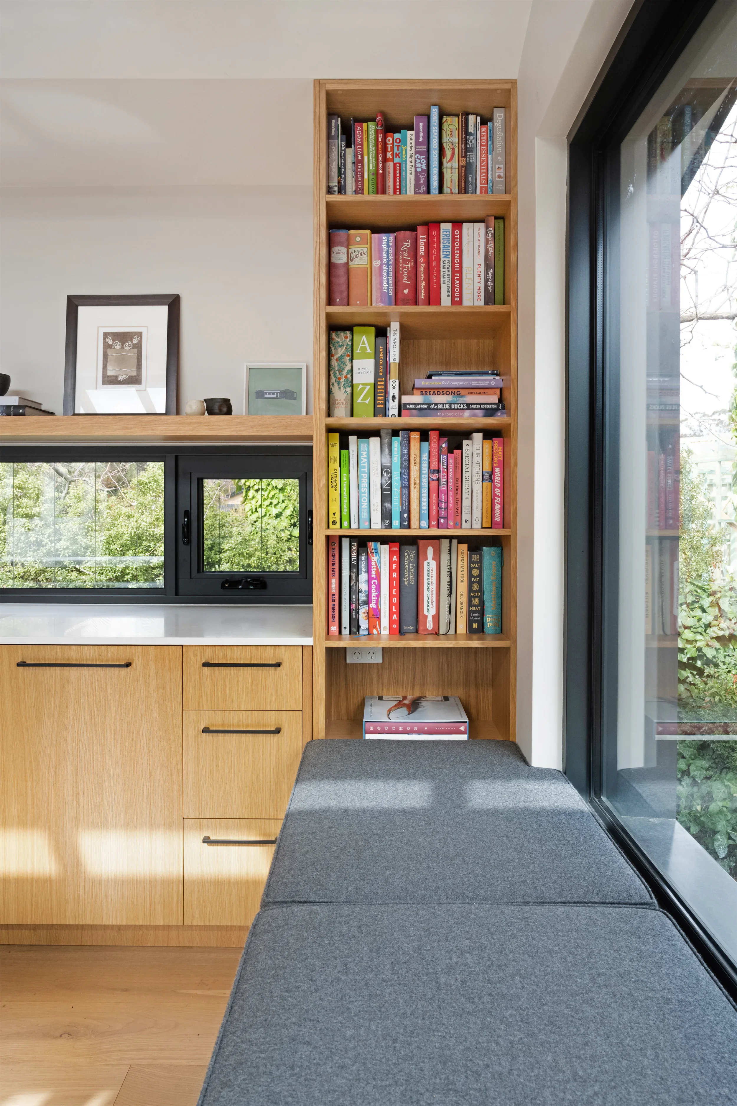 View of a modern interior with a wooden cabinet, bookshelf filled with colorful books, a window with a black frame, and part of a reception or sitting area with a gray cushioned bench.