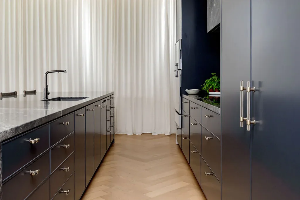 Modern kitchen with black cabinets, marble countertop, stainless steel handles, and a black sink with a faucet. White curtain in the background, wooden herringbone floor, and some green plants on the counter.