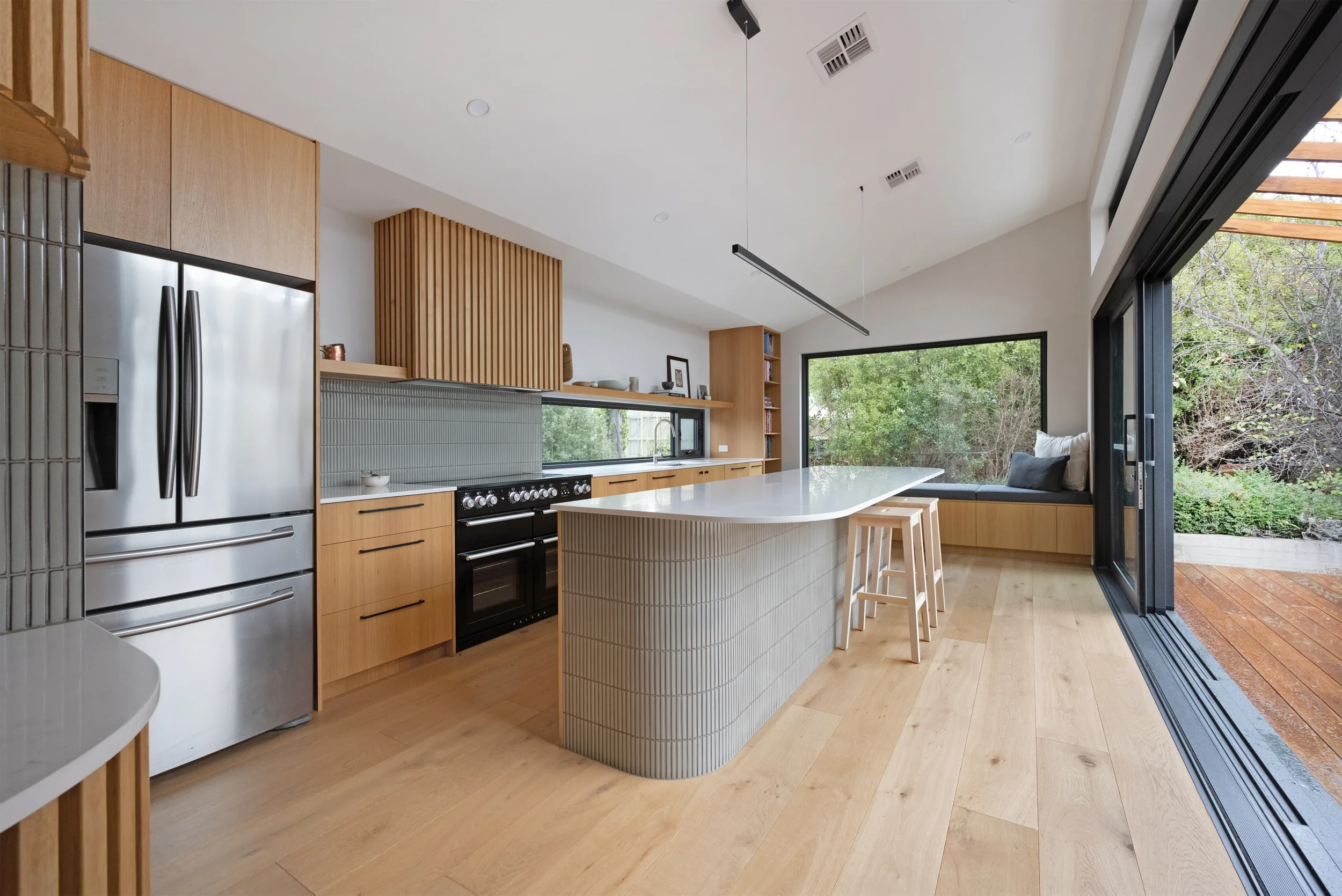 Modern kitchen with light wood cabinets, black appliances, white island with textured panels, large window with outdoor view, sliding glass door leading to patio, ceiling with black linear pendant light, and wooden floor.