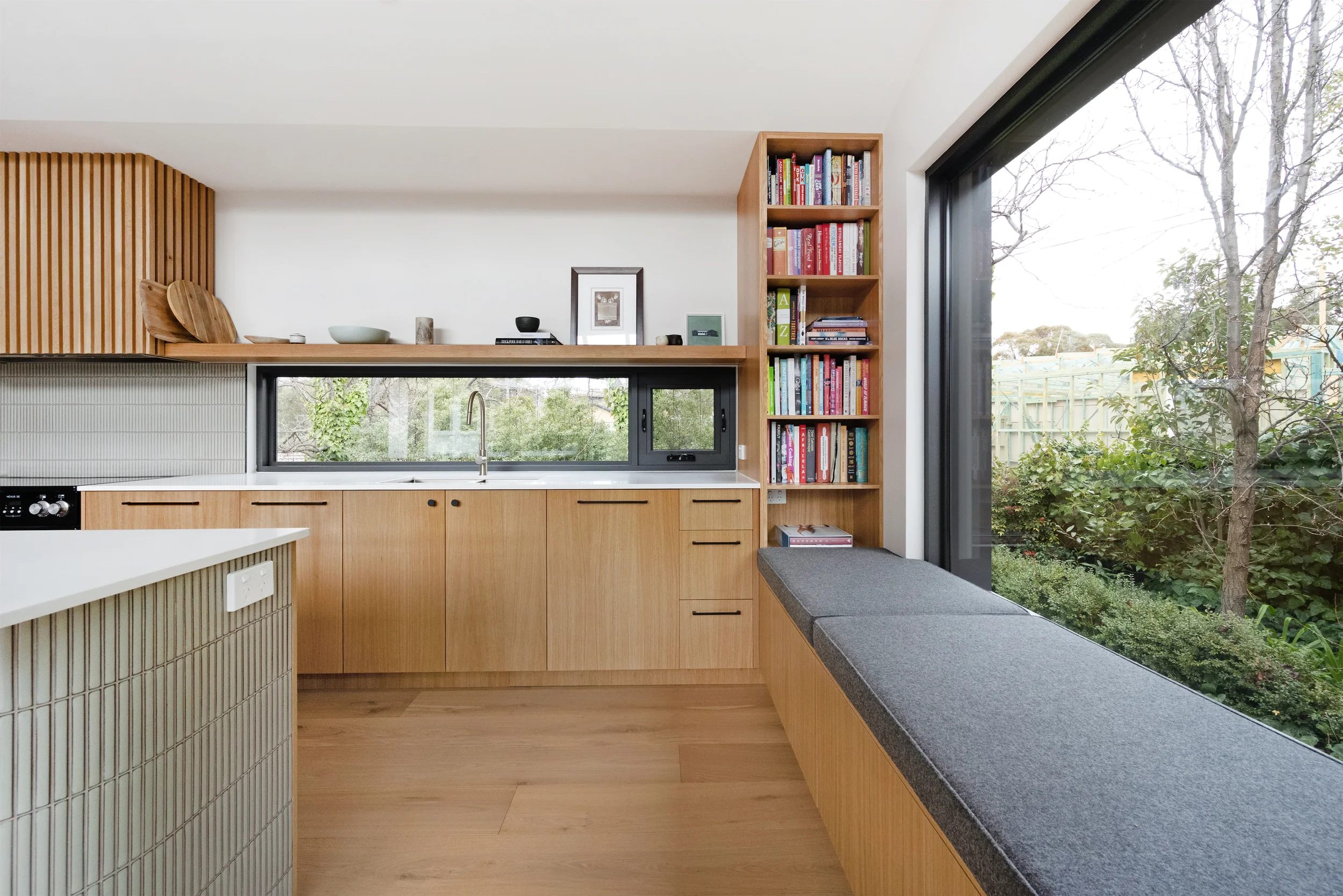Modern kitchen with wooden cabinets, a window with a view of trees, and a bookshelf filled with colorful books.