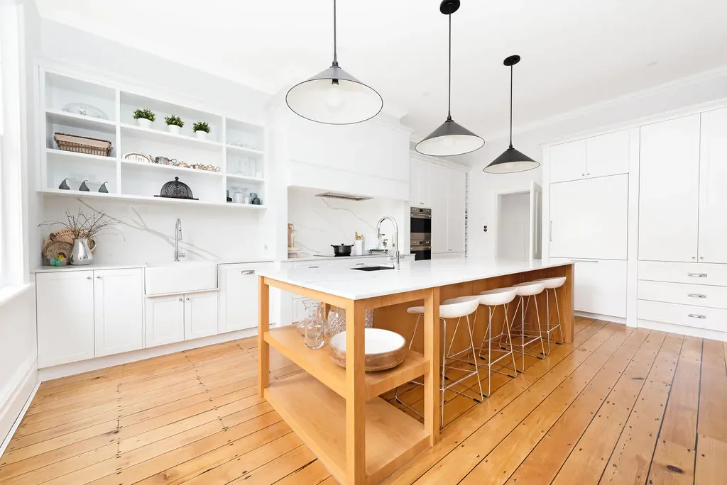 Modern kitchen with white cabinets, open shelving, a large island with a wooden base and white countertop, pendant lights, and four white bar stools. Hardwood floors and minimal decor with plants and kitchenware.