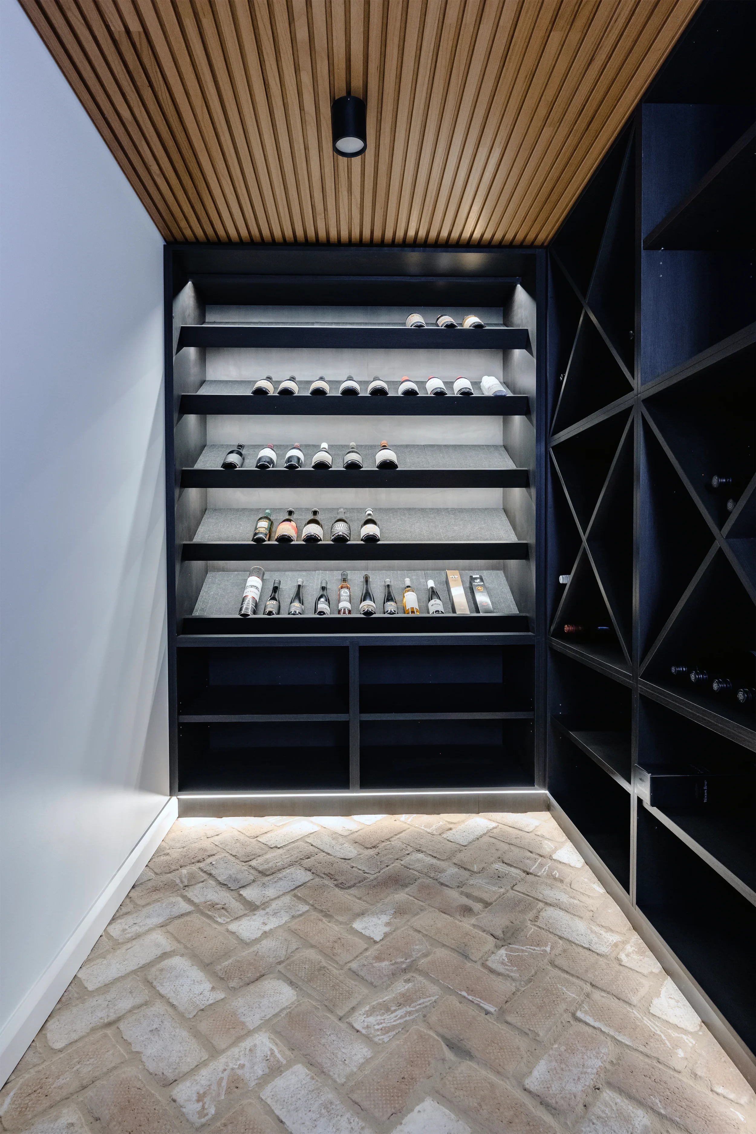 A wine storage unit built into a black wooden shelf with multiple horizontal shelves holding bottles of wine in a modern room with a ceiling of wood slats and a black ceiling light. The room has a stone floor and white walls.