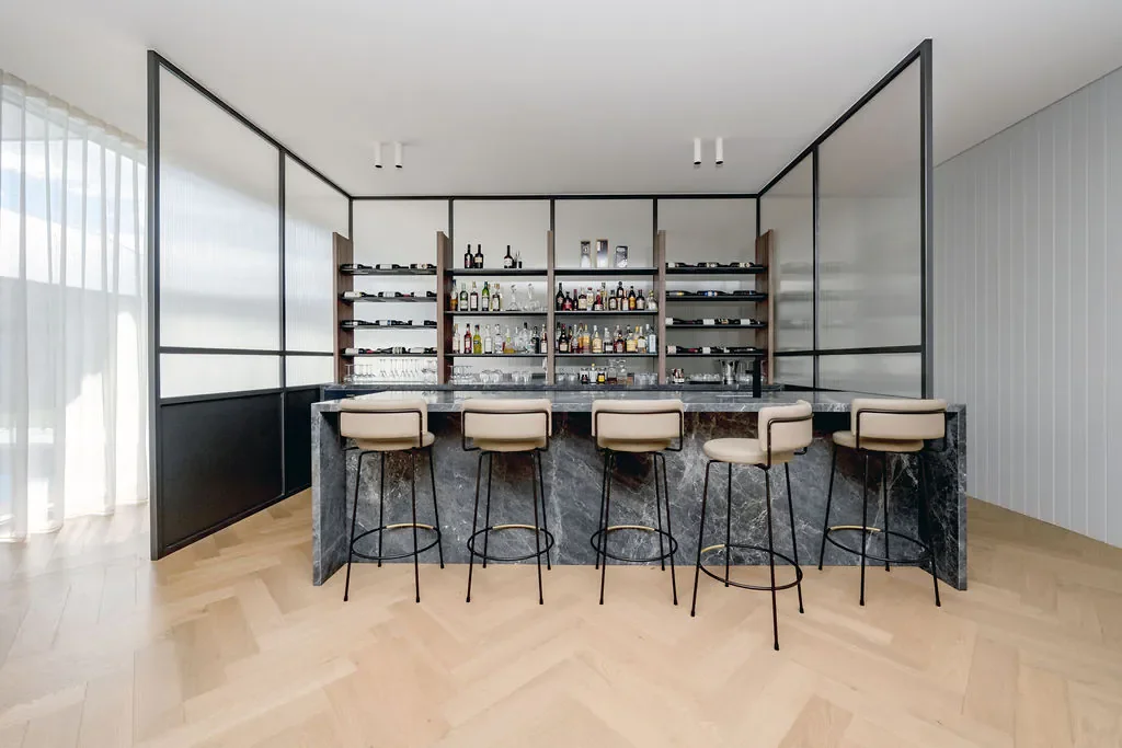 Modern home bar with a marble countertop, beige bar stools, and glass shelves stocked with liquor bottles.