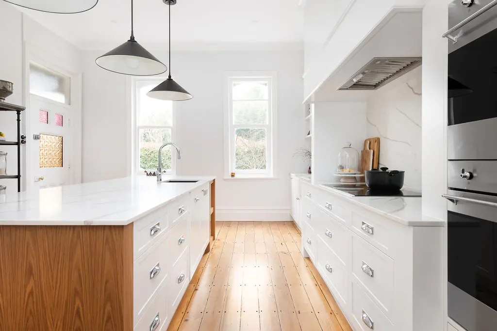 Bright, minimalist kitchen with white cabinets, a large white countertop island, light hardwood flooring, and three hanging pendant lights. Large windows let in natural light.