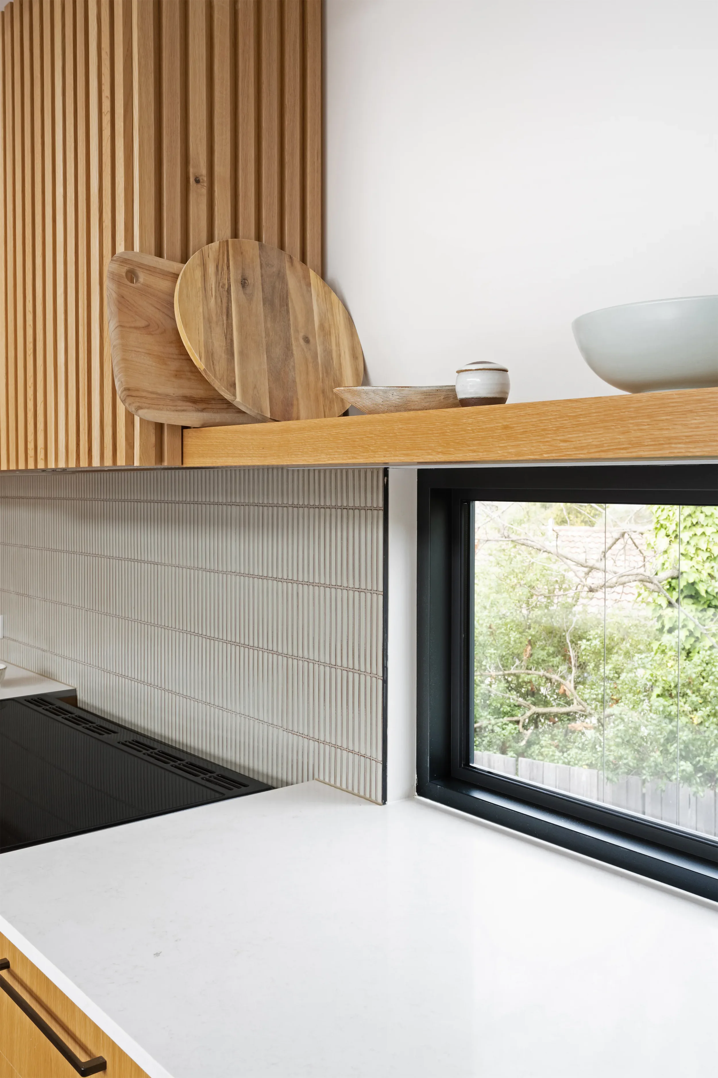 Close-up of a modern kitchen countertop with a window showing outdoor trees, and decorative wooden kitchenware including a round cutting board and a small bowl.
