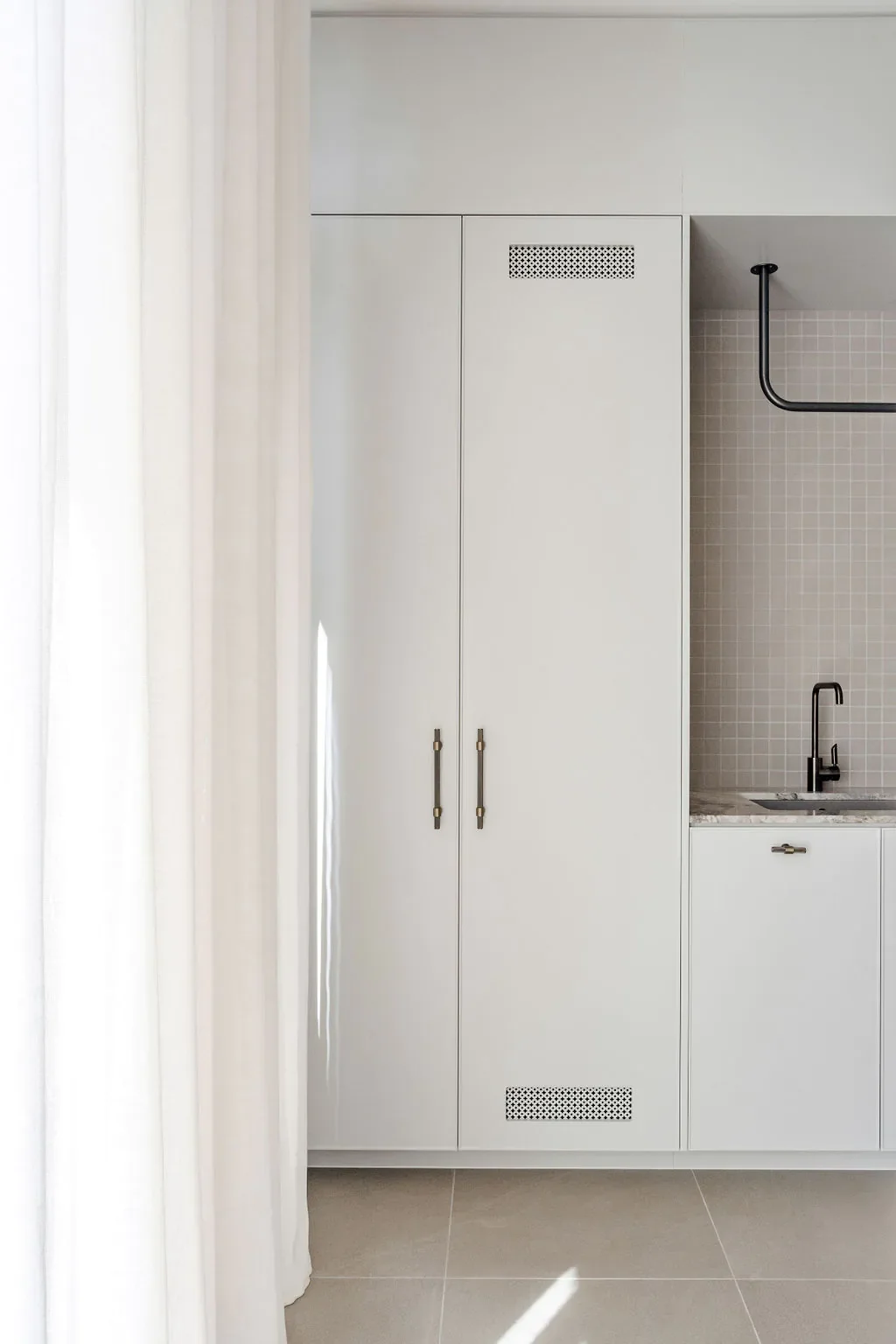 A white cabinet with two doors and gold handles next to a small kitchen sink with black faucet, beige tiled wall, and a black pipe above.