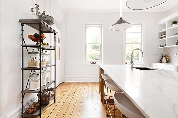 Bright kitchen with white walls, large windows, marble island, and open shelving, featuring a black metal shelving unit with glass jars and decorative items.
