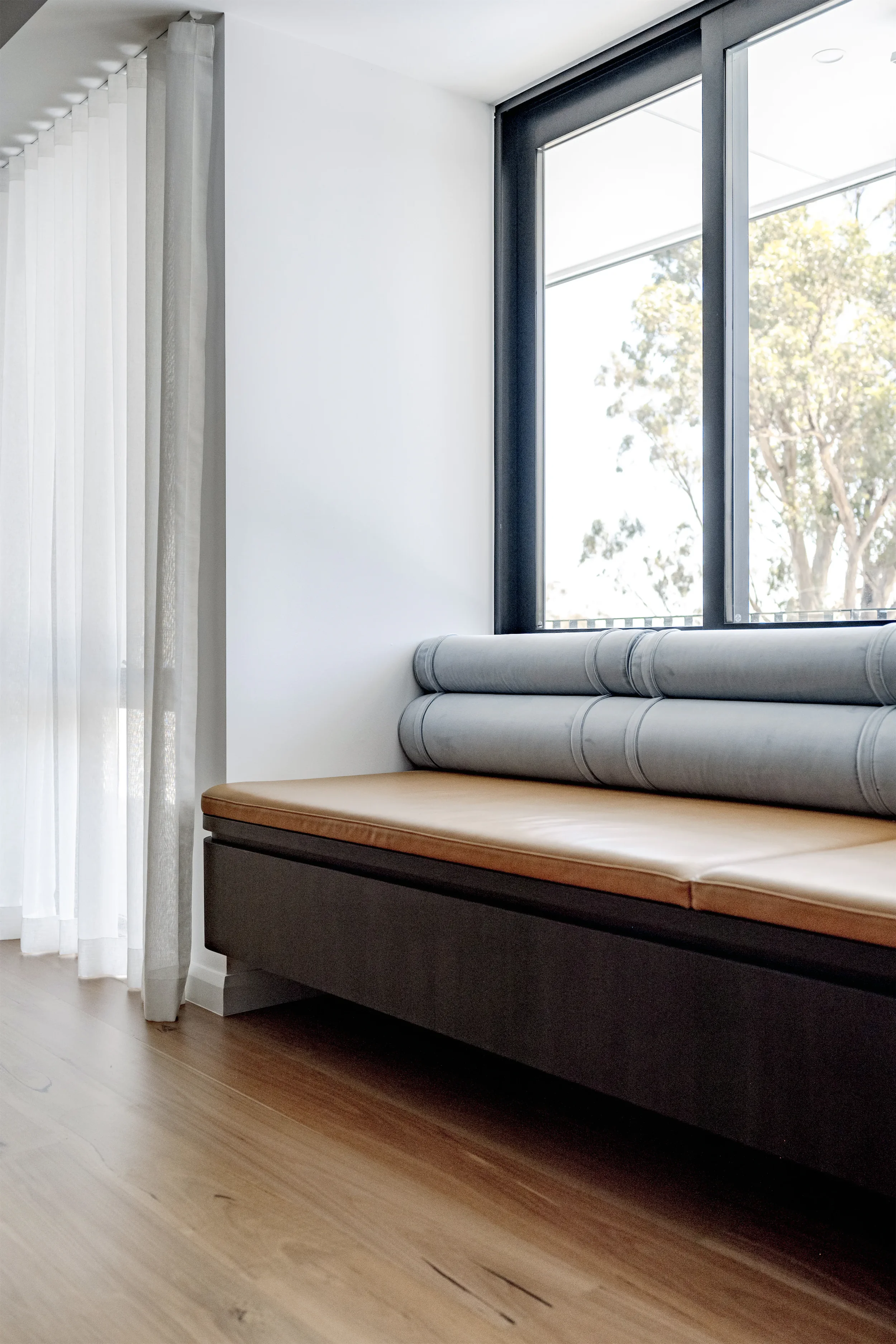 Interior view of a room with a large window, white walls, a beige cushioned bench, and light wood flooring.