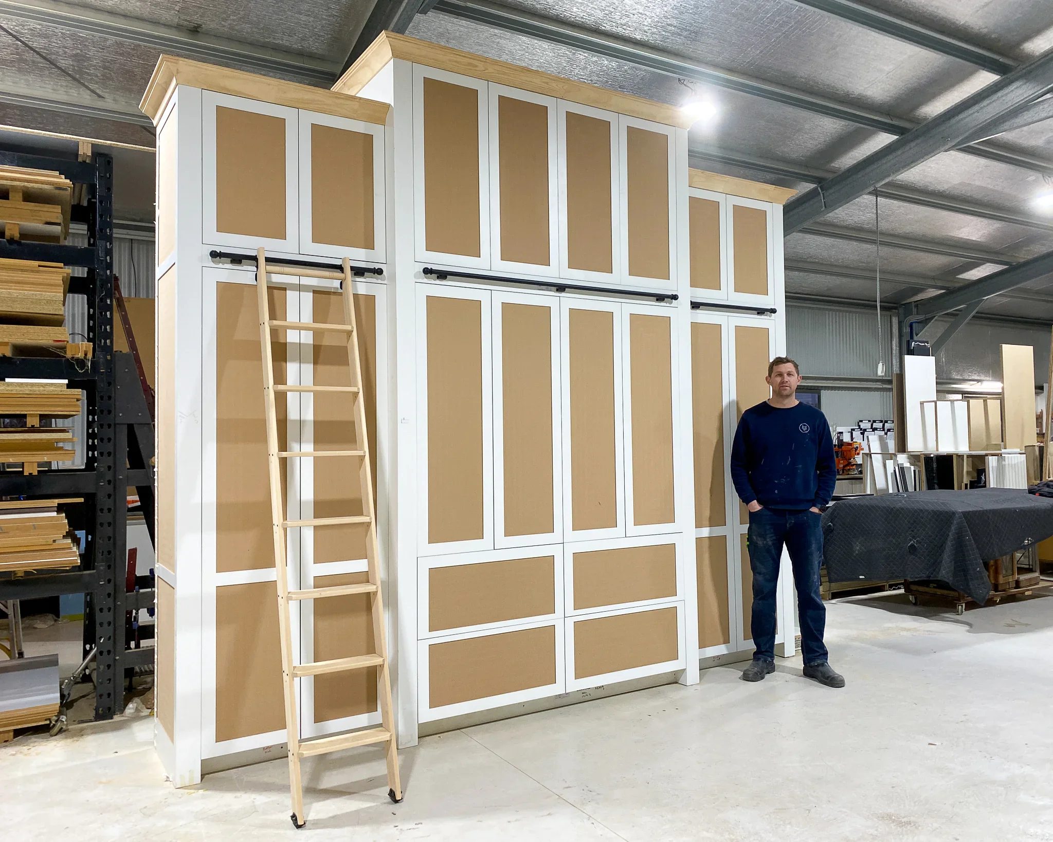 A man standing in a warehouse or workshop near a large, partially assembled piece of furniture or cabinetry with a ladder leaning against it, surrounded by various woodworking supplies and equipment.