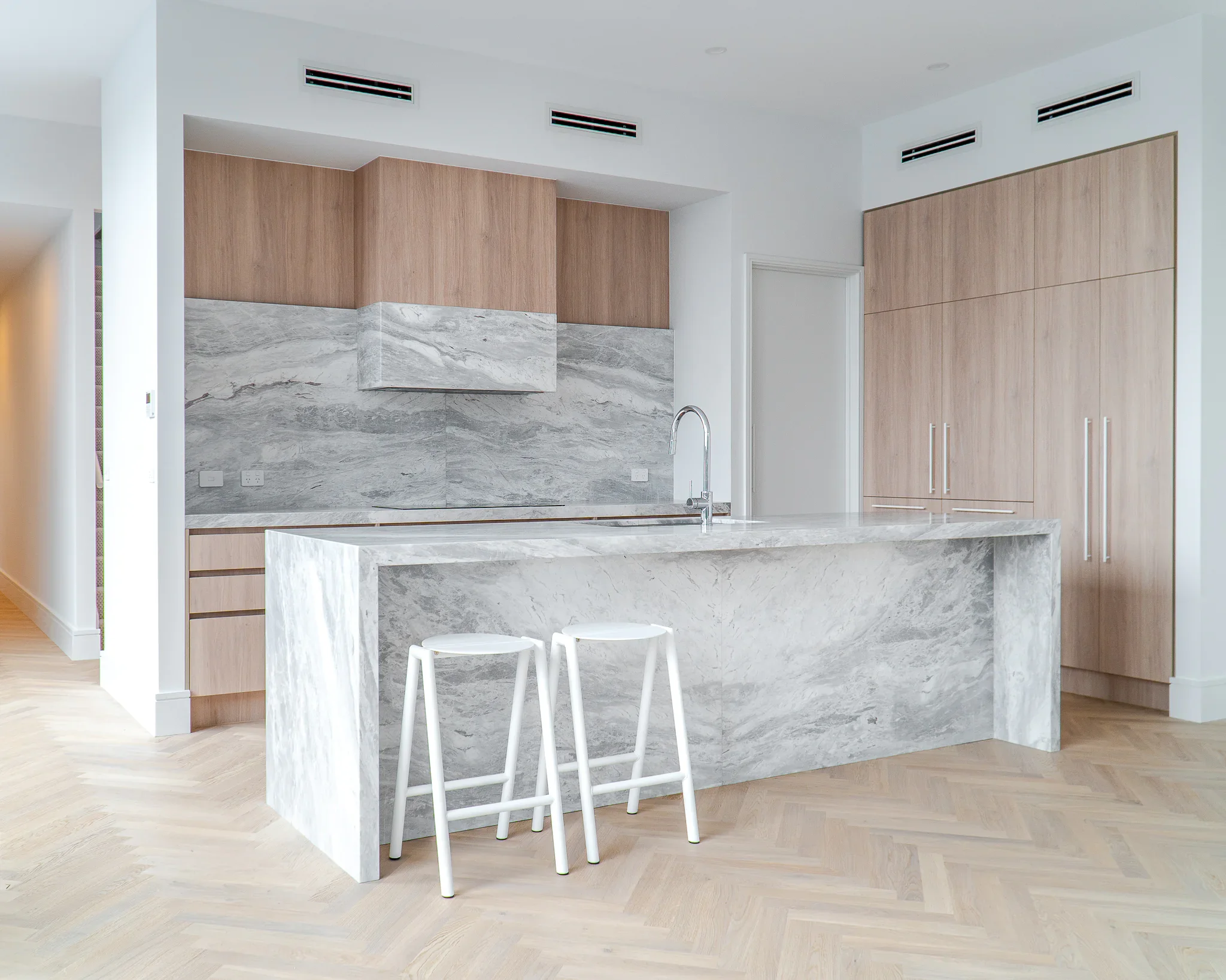 Modern kitchen with a marble island, light wood cabinetry, and stool seating, with ventilation vents on the ceiling.
