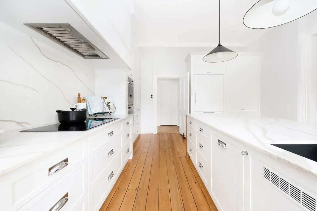 White kitchen with marble countertops, white cabinetry, wooden floor, black stovetop, and a black pot, with modern lighting fixtures.