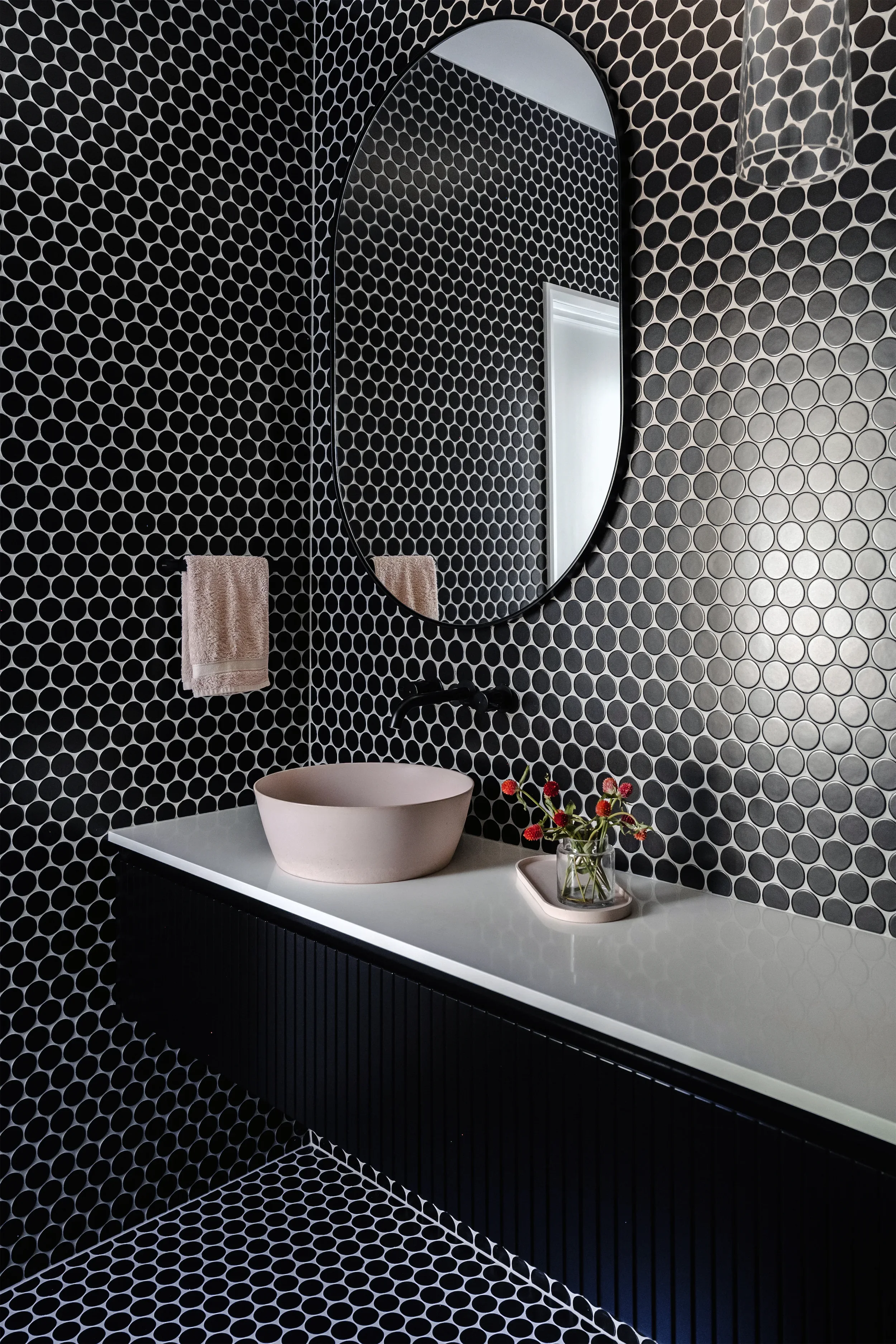 A modern bathroom featuring black and white patterned tiles on the walls and floor, a white countertop with a beige vessel sink, a round mirror, a beige towel hanging on a hook, a small vase with red flowers, and a glass pendant light.