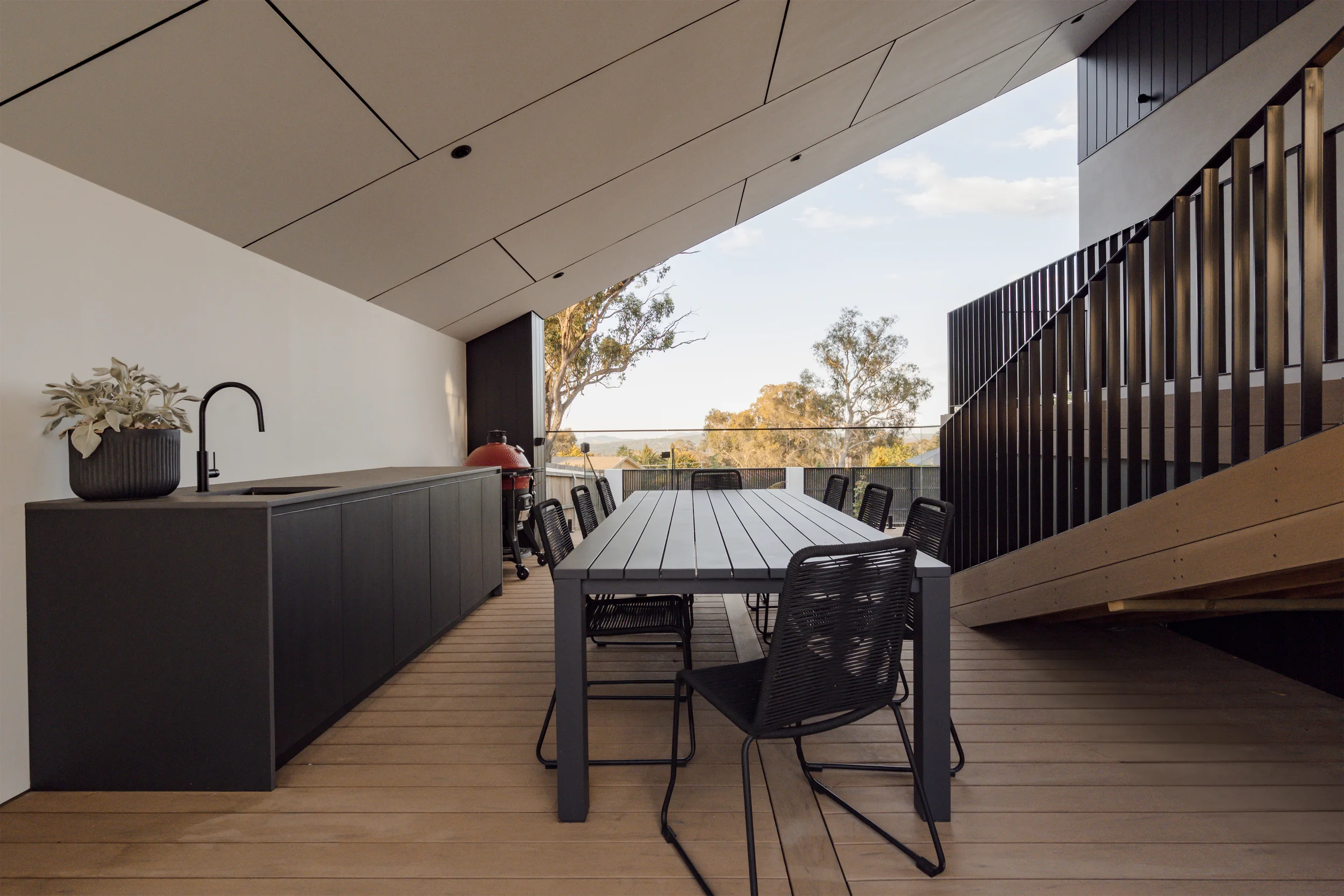 Modern outdoor balcony with a dining table and black metal chairs, black cabinetry with plant and faucet, and a view of trees and blue sky.