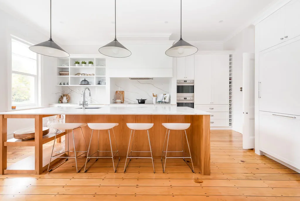 Bright, modern kitchen with white cabinets and a large white marble island with a wood base. Four white bar stools are at the island. Pendant lights hang above the island. Wooden flooring and open shelving with small plants and decorative items are visible.