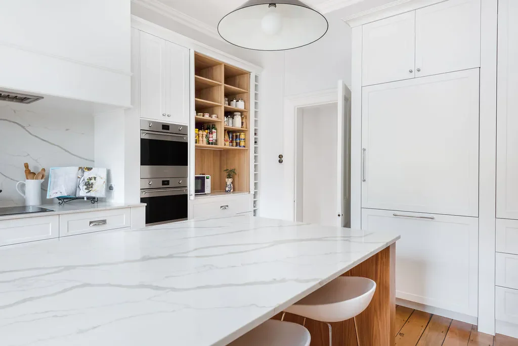 Modern white kitchen with marble countertops, built-in oven, open wooden shelves, and a pendant light fixture.