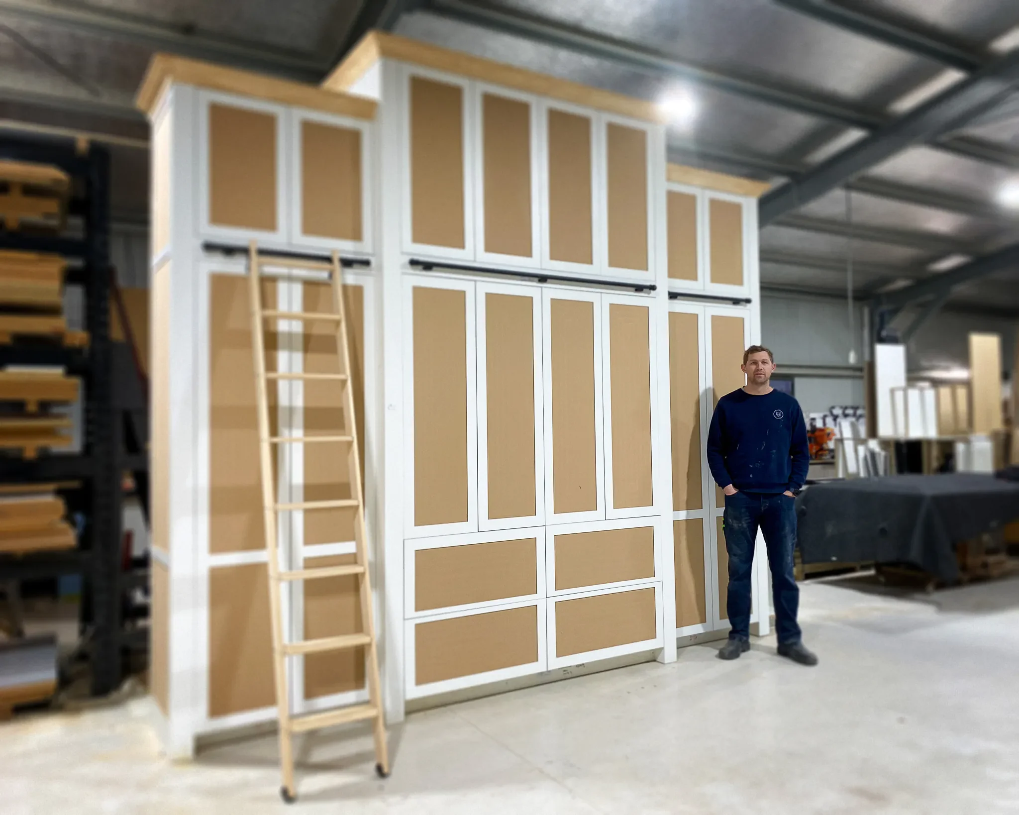 A man stands inside a warehouse near a tall, multi-compartment storage unit with a ladder leaning against it. The warehouse has a metal ceiling and shelves with various materials.